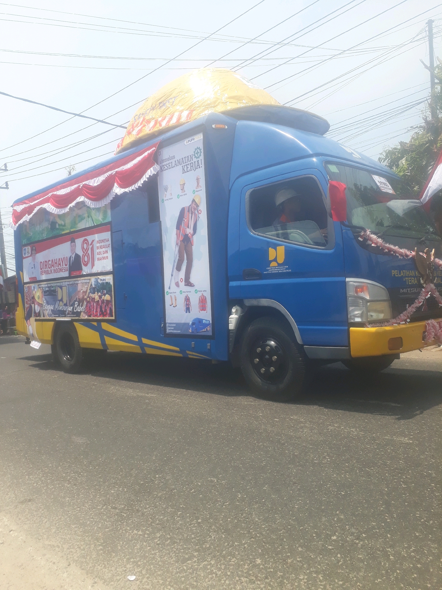 A blue truck with yellow accents is parked on a paved road. The truck is decorated with banners and posters. One poster displays "DIRGAHAYU REPUBLIK INDONESIA" and features images of two men in suits. Another poster illustrates safety guidelines for workers, including images of gloves, a hard hat, a vest, and a person with crutches. The truck also has a large yellow tarp secured to its roof. The sky is bright and hazy, suggesting a sunny day. The location is identified as the city of Pangkalpinang, Indonesia.