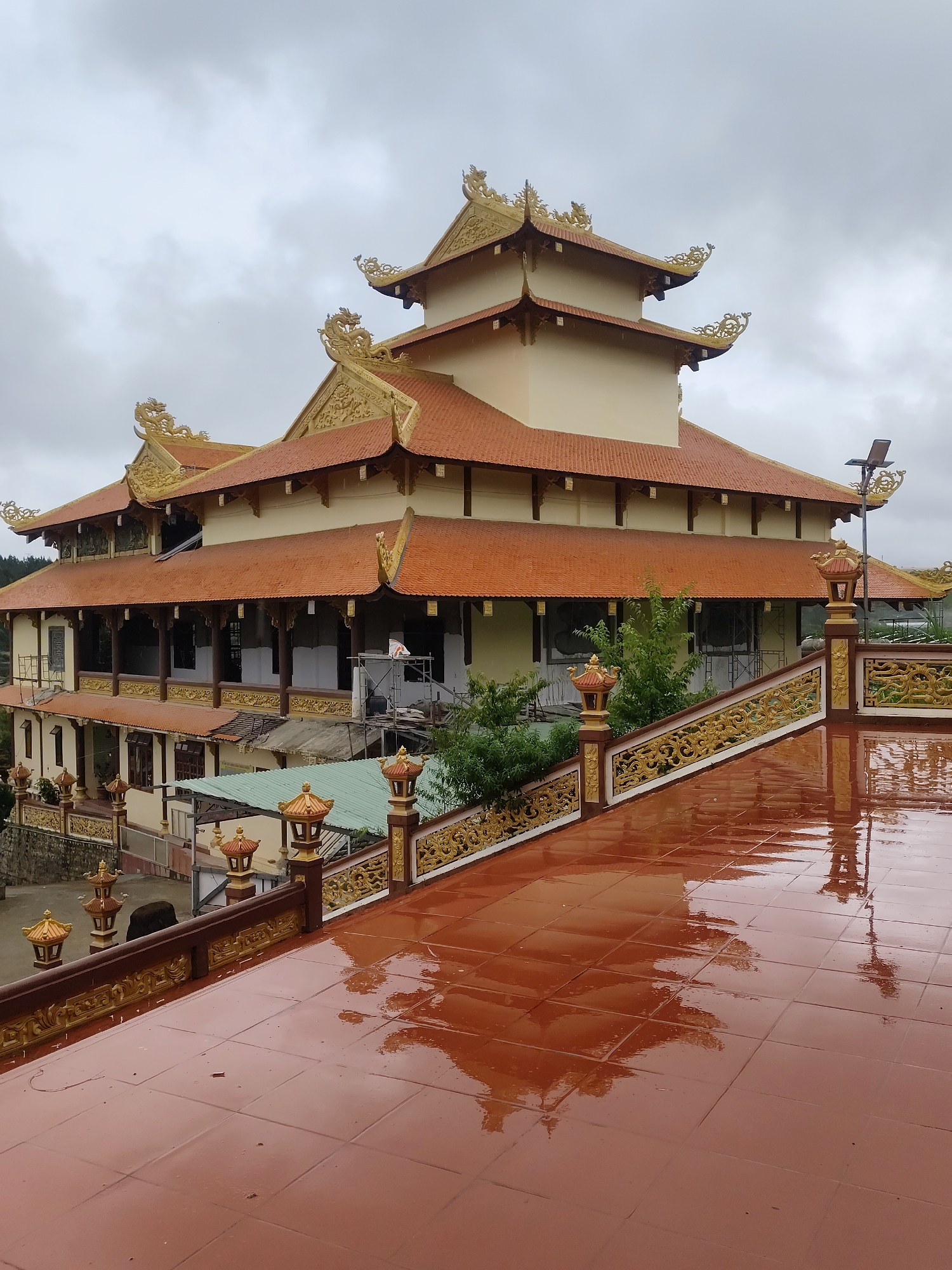 The image shows a multi-tiered Buddhist temple with traditional East Asian architectural elements, including curved, orange-tiled roofs with ornate golden dragon decorations at the eaves and corners. The temple's walls are painted a light cream color, and there are verandas with wooden columns and decorative gold railings. In the foreground, a wet, reddish-brown tiled terrace reflects the temple and the overcast sky. Scattered along the terrace and on the temple's lower levels are decorative lamps. Lush green trees and foliage are visible around the temple structure. Construction scaffolding is present on one of the temple's upper levels. The overall scene suggests a temple located in a hilly or mountainous area, under a cloudy sky, potentially after rain given the wet terrace. This structure is likely located in Lạc Dương, Vietnam, a region known for its Buddhist temples and pagodas.