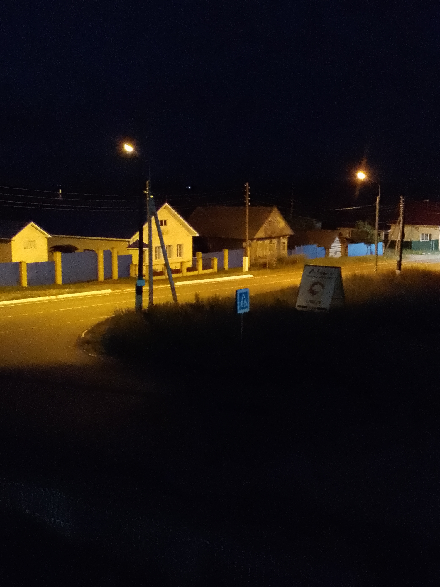 The image captures a dimly lit nighttime scene in the city of Morgueshi, Russia. The primary focus is a street illuminated by a streetlamp, casting an orange glow on the asphalt road and the surrounding grassy area. Several residential buildings are visible, characterized by their light-colored facades and pitched roofs. A blue fence with yellow posts separates the road from the properties.
In the foreground, a black A-frame sign stands on the grassy shoulder of the road. Partially visible on the sign is a logo with text, but it is not legible enough to decipher. Adjacent to the sign is a blue and white pedestrian crossing sign.
The overall ambiance is quiet and serene, typical of a rural or suburban setting at night. The lack of visible people or vehicles suggests a peaceful, uneventful moment. The environment is characterized by the contrast between the artificial light sources and the deep darkness of the night sky.