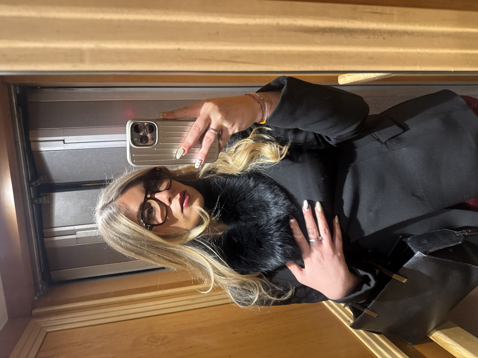 A person with blonde hair and glasses poses for a selfie. They are wearing a black coat with a fur collar and holding an iPhone in a ridged, silver-colored case. Rings are visible on their fingers. A black purse hangs from their shoulder. The photo appears to be taken in front of a mirrored wall with a wooden frame.