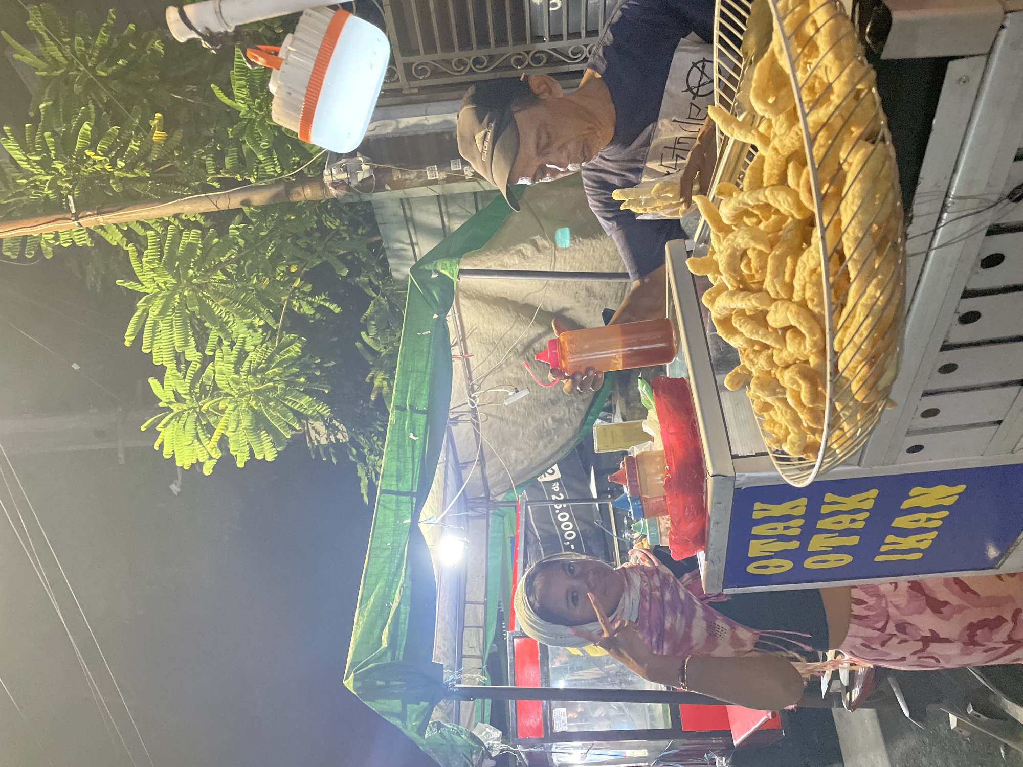 The image captures a nighttime street food scene in Central Jakarta, Indonesia. A vendor wearing a dark-colored shirt and baseball cap is preparing a dish made of fried Otak Otak Ikan (fish cake) at his mobile food stall. The fried fish cakes are piled high in a metal mesh container. A variety of sauce bottles are arranged on the cart's surface. A customer, a girl with a headscarf and floral top, poses with a peace sign. The stall is illuminated by a light. The banner on the cart reads "OTAK OTAK IKAN," indicating the specialty. The setting includes overhead tree branches and urban infrastructure, adding context to the street scene.