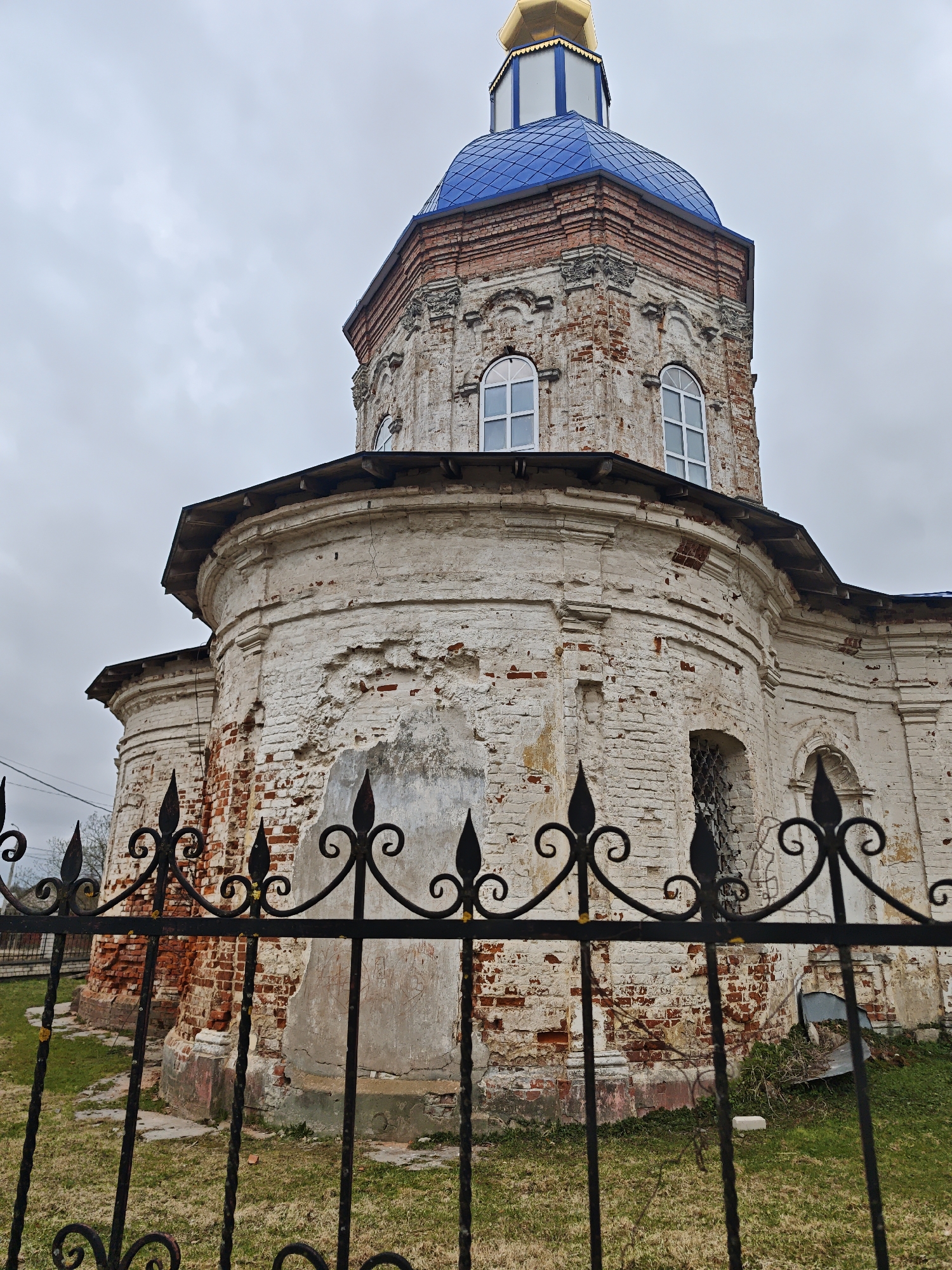 A low-angle, exterior shot depicts the architectural details of an aged church. The structure is primarily constructed of white brick, with some brick areas showing signs of deterioration and exposed red brick beneath. A black wrought-iron fence with decorative designs runs horizontally across the lower portion of the frame. The fence appears to enclose the property.
The church features a rounded base section with small, arched windows and a tiered upper section. The upper section has two arched windows with white frames and a blue, domed roof topped with a gold-colored spire. The sky is overcast, suggesting a cloudy day. The image suggests the location is in or near a rural area, with grass and sparse vegetation visible within the fenced area. The location is stated to be in посёлок совхоза "Буденновец", Russia. No people are visible in the frame.