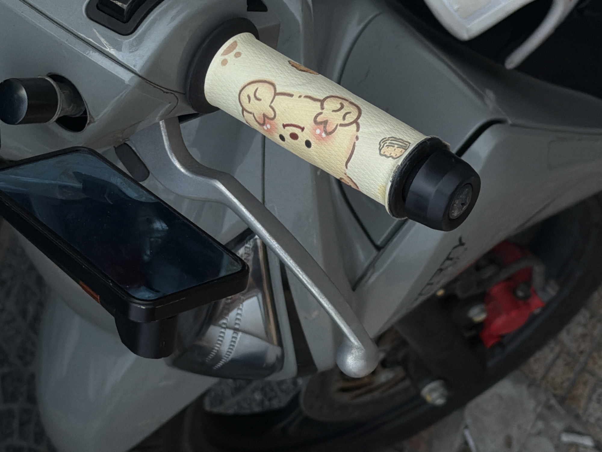 A close-up captures the handlebars of a silver scooter, possibly a motorbike, with a smartphone mounted to the left. The right handlebar is wrapped in a beige cover adorned with cartoon bear illustrations. The silver brake lever is positioned below the handlebar. Visible behind the scooter's body is a section of the front wheel, showing the black tire and a glimpse of the red brake caliper. The scooter is located in Đà Nẵng, Vietnam. The background shows a paved surface.