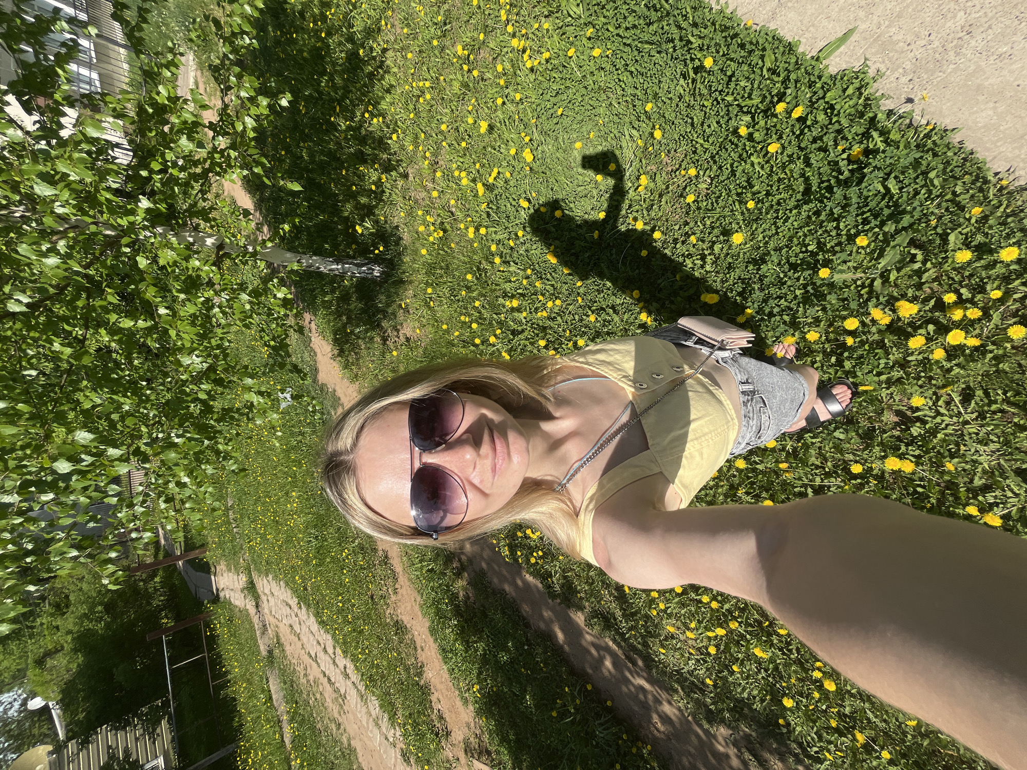 The image shows a person taking a selfie outdoors on a sunny day. The person is a fair-skinned woman with long blonde hair and sunglasses. She is wearing a yellow top and grey shorts. She is standing on grass covered in yellow dandelions. Her shadow is cast on the grass. A dirt path runs through the grass in the background. There is a birch tree on the left side of the image. The location may be Chaikovsky, Russia.