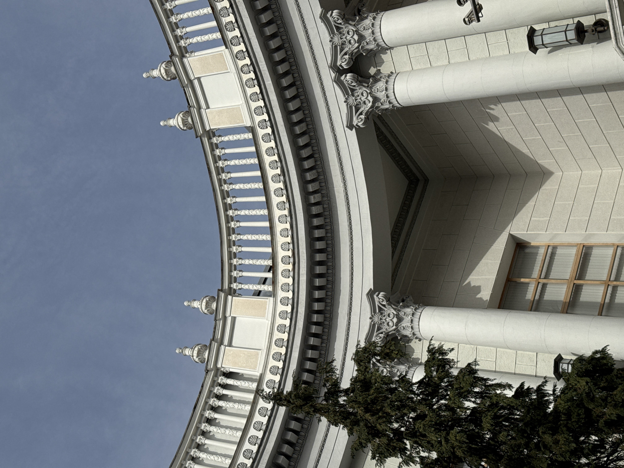 The image shows a section of an ornate, classical-style building in Sochi, Russia. The building has a curved facade with multiple architectural details, including balustrades, decorative moldings, and columns with Corinthian capitals. A tree obscures the lower portion of the facade, while the upper section of the building features windows and exterior lighting fixtures. The background consists of a clear, blue sky. The architecture suggests a public or monumental building.