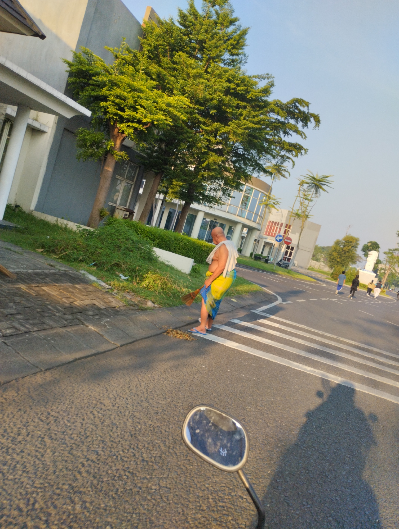 A man in a sarong and a white cloth draped over his shoulders sweeps leaves on a sidewalk next to a road with a pedestrian crossing. The scene appears to be in the city of Sidodadi, Indonesia, as indicated by the building architecture, lush greenery, and what appears to be a zebra crossing common in many Indonesian cities. The man is bald and appears to be middle-aged or older. He is using a broom made of natural fibers. In the background, several people are walking across the zebra crossing. The sun is low in the sky, casting long shadows. A motorcycle mirror and its rider's shadow are visible in the foreground. The environment includes a modern building with large glass windows, trees, and well-maintained landscaping.