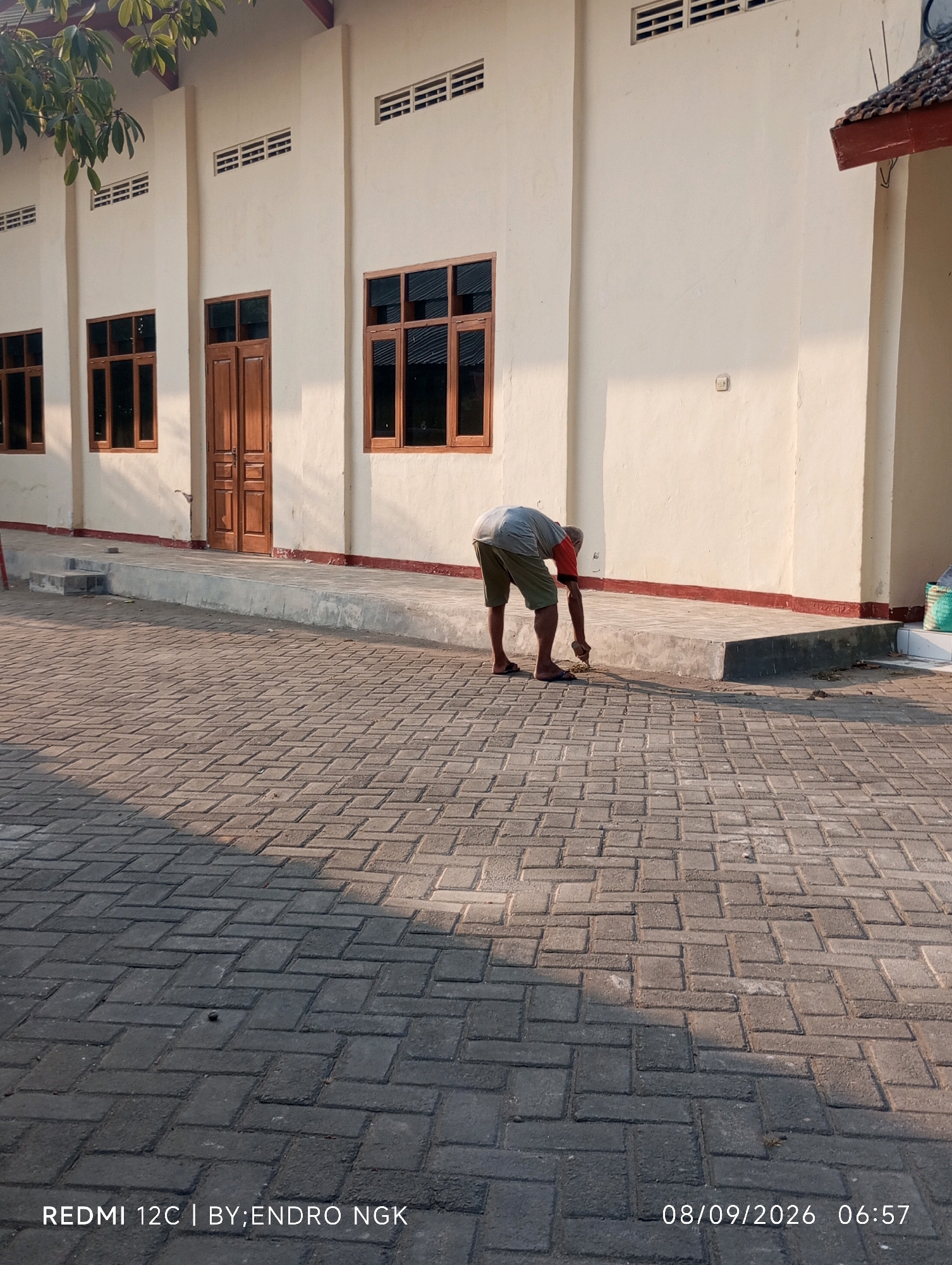 In the city of Guyangan, Indonesia, an elderly man in a grey t-shirt and green shorts is bent over, sweeping debris from a paved walkway in front of a light-colored building. The building has several windows with dark frames and a prominent wooden double door. A small set of concrete stairs leads up to the building's entrance. The walkway is paved with interlocking bricks, creating a geometric pattern. The scene is illuminated by the warm, low light of what appears to be sunrise or sunset, casting long shadows across the paved area. A few branches of a leafy tree are visible in the upper left corner. The lower right corner of the image displays a timestamp: "08/09/2026 06:57" and the text "REDMI 12C | BY;ENDRO NGK".