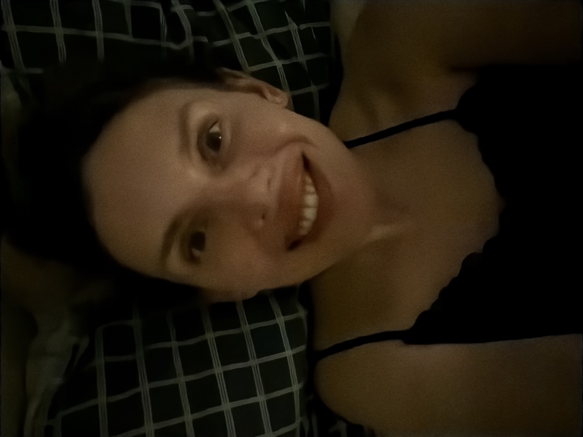 A woman is lying on her side on a checkered pillow or cushion, smiling towards the camera. She is wearing a dark spaghetti-strap top. The lighting is dim, suggesting an indoor environment, possibly a bedroom. The image does not contain enough information to confirm its location in Budva, Montenegro.