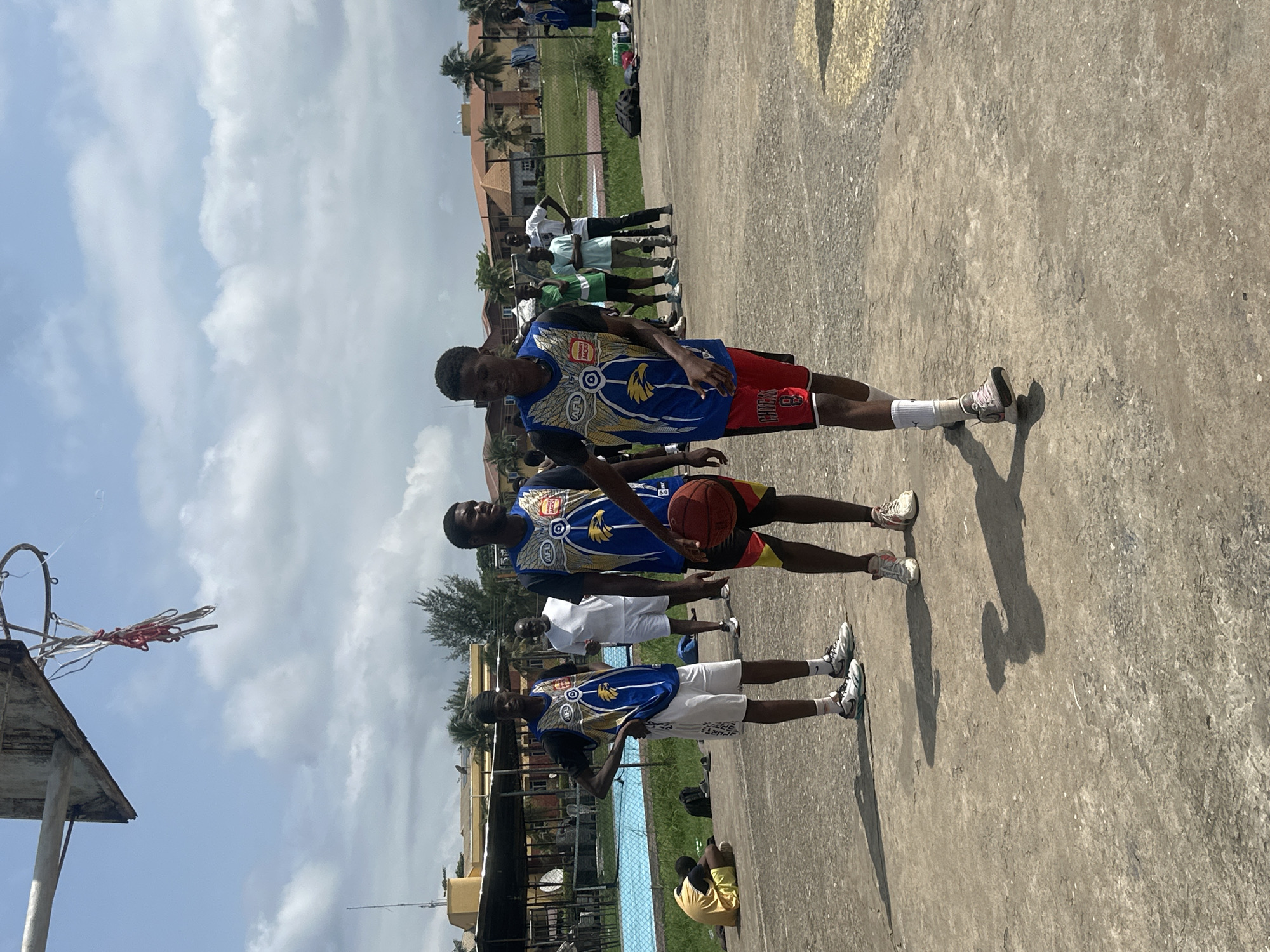 In Ajara, Nigeria, a group of young men are playing basketball on an outdoor court under a cloudy sky. In the foreground, a young man wearing a blue and yellow jersey with "AFL" and an eagle logo, red shorts, and white socks is dribbling a basketball. To his left, another young man in a similar jersey and white shorts stands with his hands on his hips, looking away. Further in the background, other individuals are visible on the court, some in athletic wear and others in casual clothing. A swimming pool and buildings are situated behind a fence to the left of the court. The court itself is made of concrete, with some patchy areas and visible shadows cast by the players and the basketball hoop. The basketball hoop has a weathered backboard and a net made of red and white ropes.