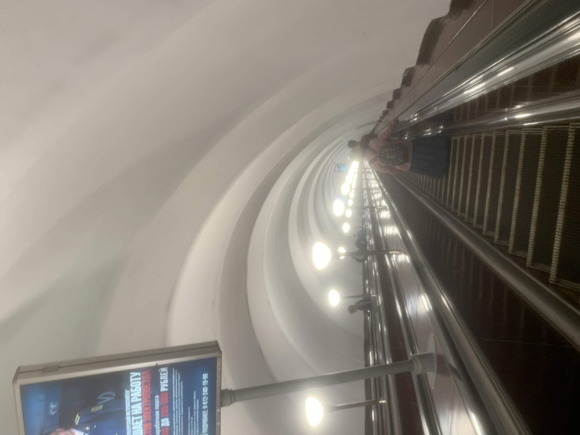 The image captures a deep, long escalator tunnel within a metro station, viewed from a perspective looking down the escalator. The tunnel features a high, arched ceiling with subtle ribbed architectural details, painted in a light, neutral color, creating a sense of immense depth.
On the right side of the frame, a continuous escalator descends, characterized by its dark, segmented steps and bright, metallic handrails. Several indistinct figures are visible on the escalator, appearing to be passengers commuting. One person in the foreground, wearing dark attire, stands on the escalator.
A series of bright, round lights are mounted along the right wall of the tunnel, above the escalator, providing illumination and reflecting off the metallic surfaces. On the left side of the tunnel, a brightly illuminated rectangular advertisement board is prominently displayed. The text on the billboard is in Russian, indicating job opportunities and salary information. Clearly visible phrases include "ОПЛАЧИВАЕТ НА РАБОТУ" (Pays for work/Work pays), "ДО 205 000 РУБЛЕЙ" (Up to 205,000 RUBLES), and a phone number "8-812-240-19-99" for "ПОДРОБНЕЕ" (Details). Other partially visible text includes "зарaботная плата" (salary) and "ОТ 50 ДО 205 000" (FROM 50 TO 205,000).
The scene depicts a typical deep underground metro system, common in older Russian cities, with the artificial lighting indicating an indoor environment.