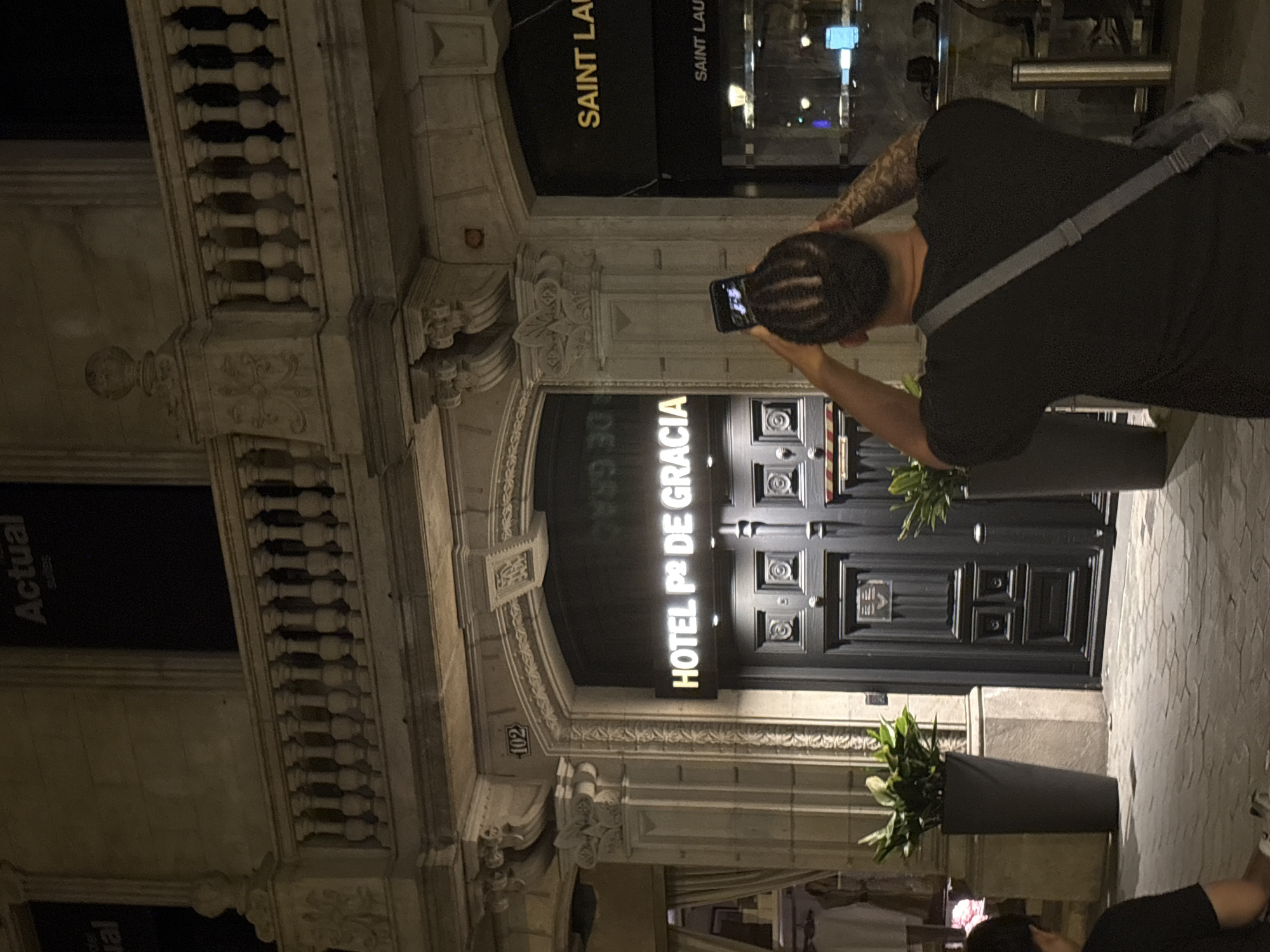 The image depicts an individual using a smartphone to photograph the entrance of Hotel Pº de Gracia in Barcelona, Spain. The hotel's name is illuminated above its dark wooden doors, which feature decorative panels. Two tall, cylindrical planters containing foliage flank the entrance. The building's façade is ornate, featuring classical architectural details. A Saint Laurent store is visible above and to the left of the hotel entrance. Additional people can be seen at the bottom of the frame, possibly waiting or passing by. The scene is lit primarily by artificial light, suggesting it is either dusk or nighttime.