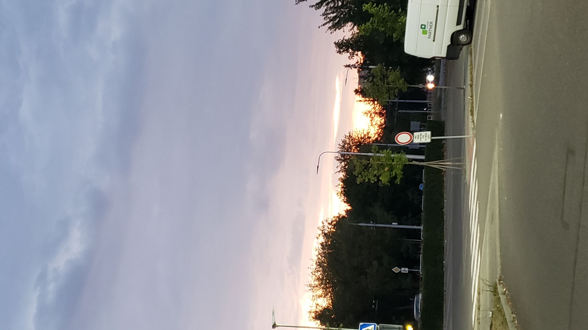 The image captures a sunset scene at a crosswalk in Prešov, Slovakia. The sky is filled with hues of purple, pink, and orange, indicating the sun is low on the horizon and partially obscured by clouds and trees. A white van with the logo "PARTNER" is parked to the right of the frame. In the foreground, a pedestrian crosswalk is visible on an asphalt road. Several streetlights and traffic signs are present in the scene, along with lush green trees lining the background. The overall atmosphere is calm and serene.