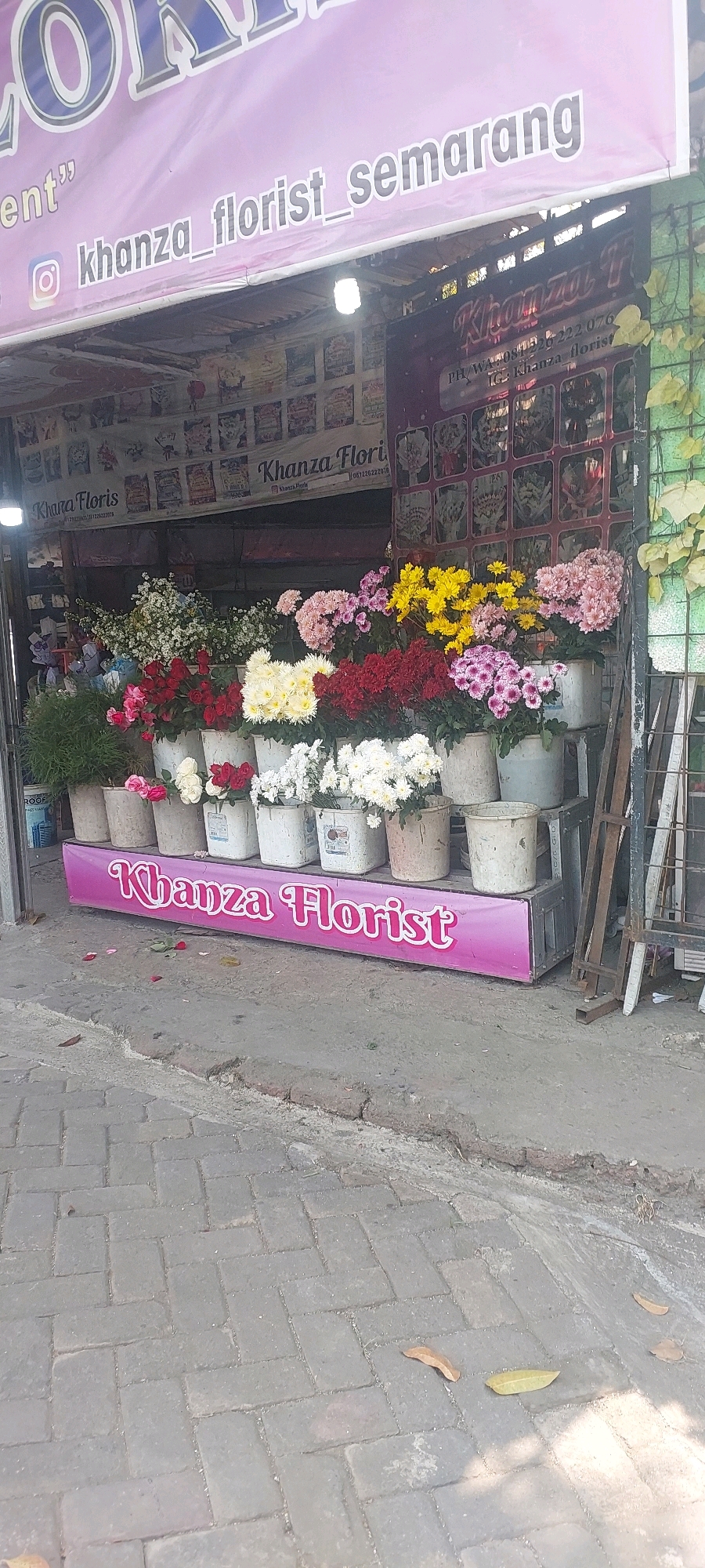 This is a low-angle, full-shot photograph of a florist shop in Semarang, Indonesia. The shop is identified by a large pink banner across the top reading "Khanza Florist Semarang" with an Instagram logo next to it. Below this, the shop itself is visible, displaying a variety of colorful flowers in buckets. The flowers include red, white, yellow, and pink chrysanthemums, as well as other greenery. A pink banner with "Khanza Florist" written in white cursive is displayed at the bottom of the shop's facade. The shop appears to be located on a sidewalk made of gray brick pavers. The overall impression is that of a small, local flower business.