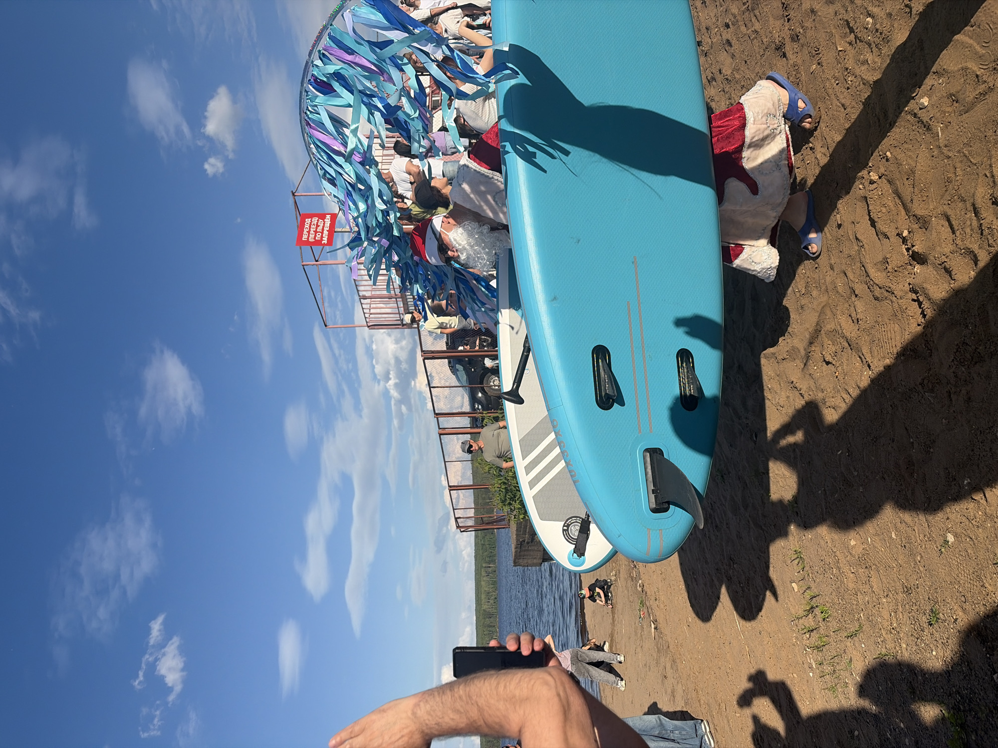The image depicts a sunny outdoor scene on a sandy beach next to a body of water, likely the Kama River in Chaikovsky, Russia. In the foreground, two large paddleboards are prominently displayed, with the closest one being a vibrant turquoise. A person is partially visible carrying the turquoise paddleboard, wearing a red and white patterned garment and blue slip-on shoes. A shadow of this person and the paddleboard is cast on the sand.
In the middle ground, a group of people are gathered. Some individuals appear to be wearing festive attire, including what looks like a Santa Claus figure with a white beard and red hat. They are positioned behind the paddleboards, and some are looking towards the water or the activity. A red sign with Cyrillic text is visible on a structure behind the group, which translates to "Crossing (passage) on ice is prohibited." This sign suggests the location is an area where ice might form, reinforcing the idea of a riverside or lakefront setting.
Further back, the water of the river or lake is visible, with a treeline on the horizon. The sky is a clear blue with scattered white clouds. Several other individuals are seen in the background, some walking or standing near a fence and a parked vehicle. The overall atmosphere appears to be one of a community event or gathering, possibly related to water sports or a festive occasion.