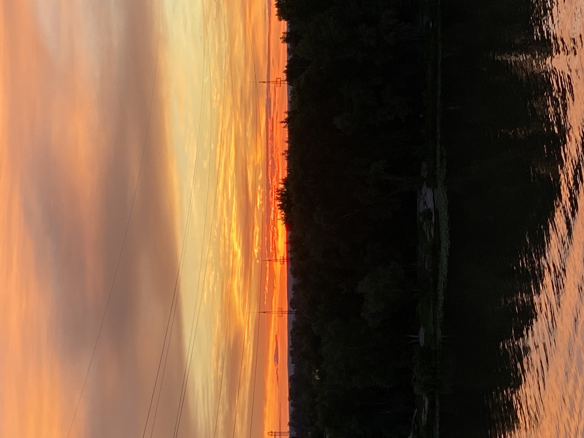 The image captures a sunset over a body of water, likely a river, in Novokuybyshevsk, Russia. The sky is ablaze with vibrant orange and yellow hues, transitioning into softer pink and purple tones as it rises. Power lines stretch horizontally across the sky, partially silhouetted against the colorful backdrop. Below the sky, a dense, dark green forest occupies the middle ground, forming a stark contrast to the bright heavens. Two tall, lattice-like towers, possibly communication or power pylons, are visible above the treeline, with one slightly to the left and another towards the center-right of the frame. The foreground is dominated by the dark surface of the water, reflecting the warm colors of the sunset, creating shimmering patterns on its surface. The overall scene is serene and natural, with the man-made structures subtly integrated into the landscape.