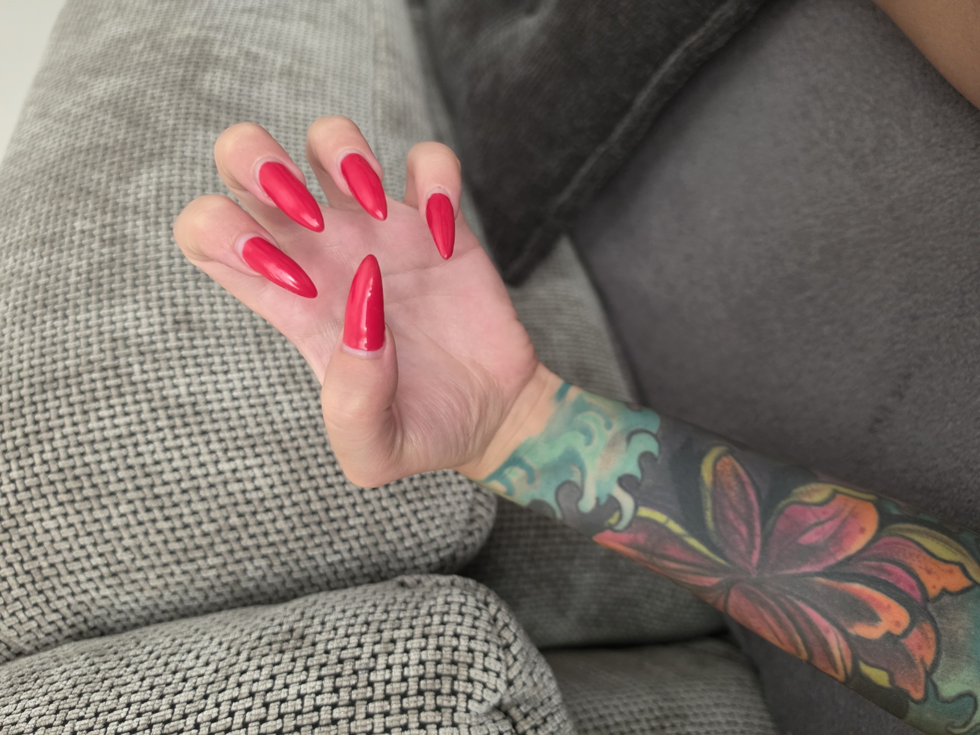 The image shows a close-up of a person's left arm and hand, resting on a textured grey couch. The hand has long, stiletto-shaped fingernails painted a vibrant red. The person's arm features a colorful tattoo that appears to depict a large flower with petals in shades of red, orange, and yellow, surrounded by swirling green and blue elements, possibly representing water or abstract shapes. The background consists of the couch, with a darker gray pillow visible to the right. The lighting suggests an indoor setting, likely during the daytime, given the brightness and clarity of the image. No text is visible.