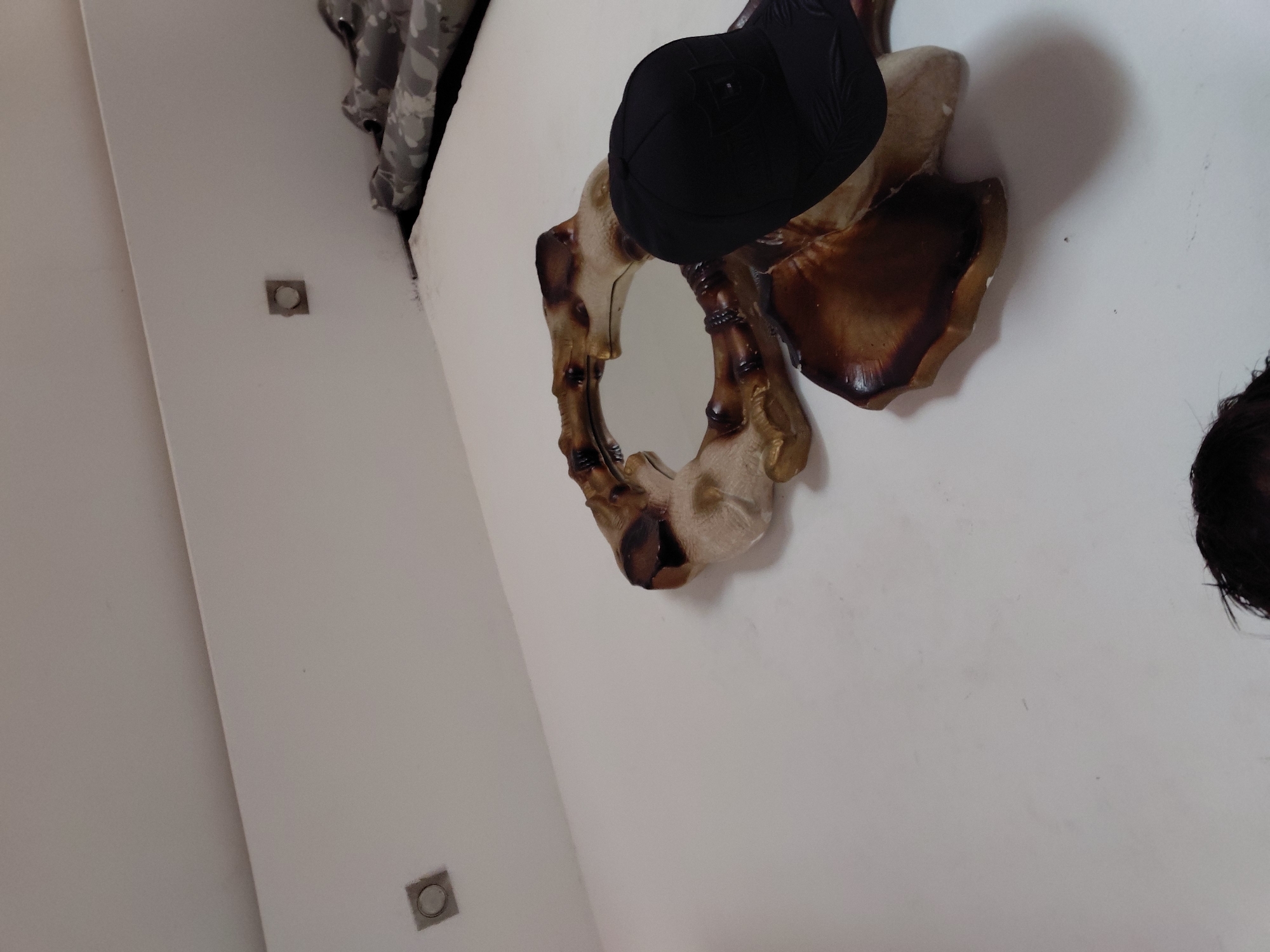 The image captures an indoor scene featuring a decorative mirror, a black hat, and architectural details on a white wall. The perspective is angled upwards, suggesting the photo was taken from below.
The central subject is an ornate wall mirror with an irregular, organic frame, possibly made of carved wood or a material resembling bamboo, finished in shades of brownish-beige and dark brown. A small, circular mirror is embedded within its unique design. A black, sporty hat, possibly a baseball cap, is placed on the upper part of the mirror's frame. Shadows cast by the mirror and hat are visible on the white wall, indicating an internal light source.
Two square, recessed light fixtures are visible on the white wall, one in the upper left and another in the lower left portion of the frame. In the upper left corner, near the ceiling, a patterned fabric, which appears to be a camouflage curtain, can be seen. A small section of dark, possibly curly hair is visible in the bottom right corner, hinting at the presence of a person close to the camera or mirror.
The setting appears to be a room, possibly a bedroom or living area. No specific activity is occurring; the image presents a static arrangement of objects. The time of day is not discernible. No text is visible in the media.