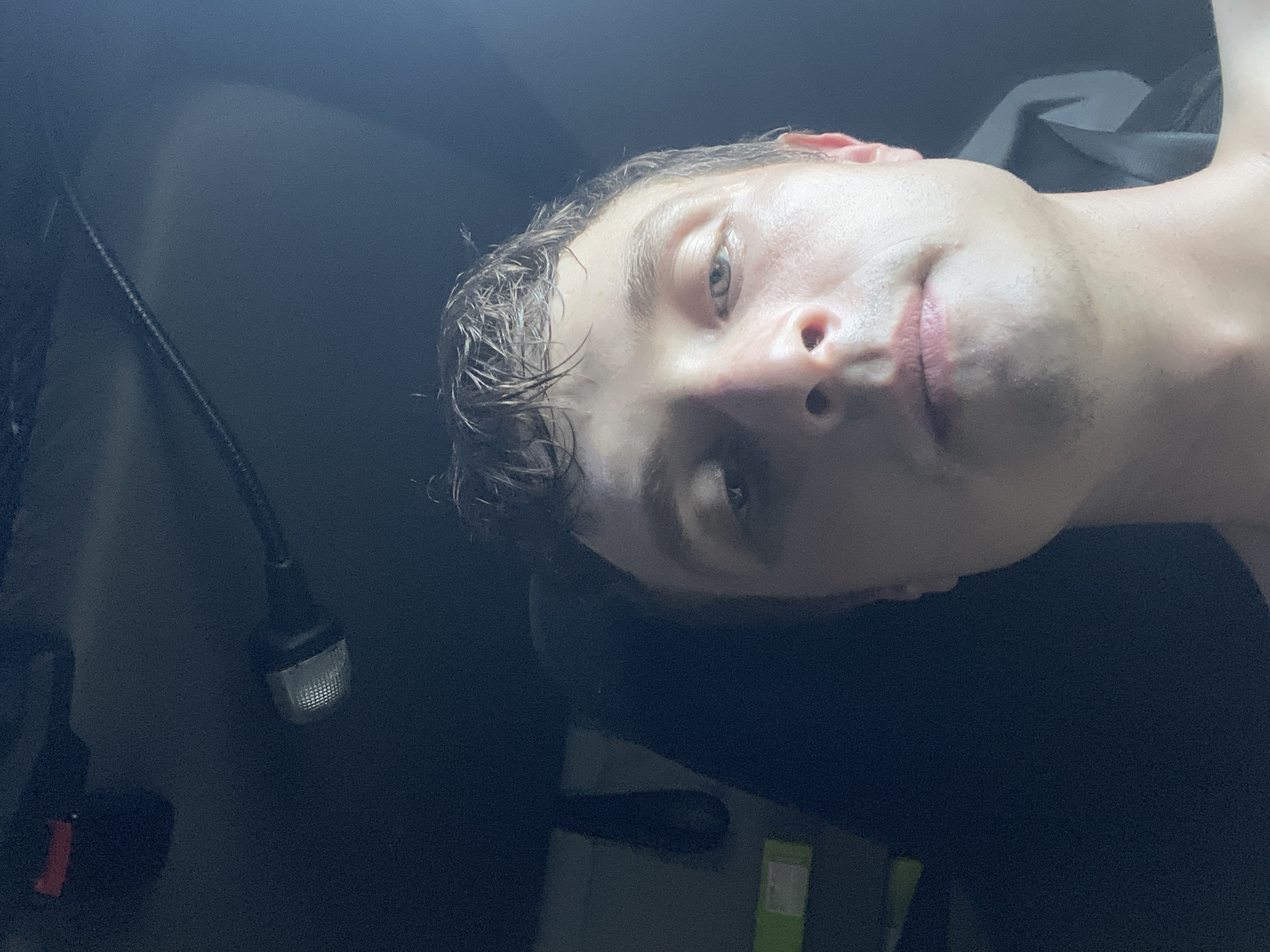 The media file is a close-up, tilted selfie of a young Caucasian person with wet hair and a pale complexion. The lighting is dim, suggesting it might be nighttime or an indoor setting with low light. The person's head is angled, with their face partially obscured by their hair. Their eyes are open, and they appear to be looking towards the camera. Their lips are slightly parted. The background is dark and appears to be the interior of a vehicle, possibly a car or truck, with a dark fabric seat or headrest visible. There's a black cable with a small, illuminated object at the end, possibly a light or accessory, visible on the left side of the frame. The overall mood is casual and personal. There is no visible text in the image.