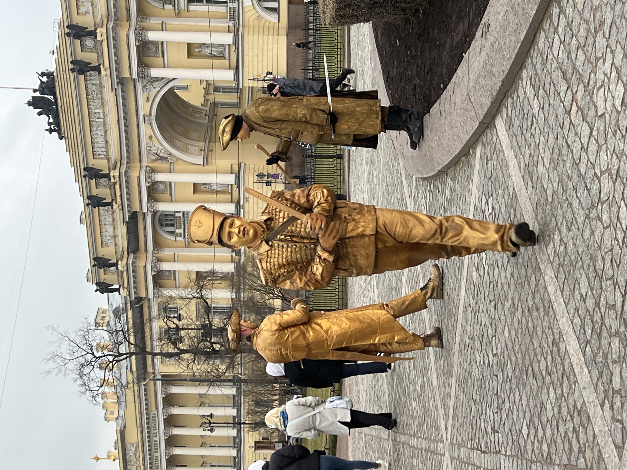 The image captures a street scene in Saint Petersburg, Russia, featuring performers in golden attire. Three performers, dressed in elaborate golden costumes reminiscent of historical military uniforms, are holding swords and standing among pedestrians. The architectural backdrop indicates a classic building with columns, statues, and decorative elements, suggesting a location near a significant landmark. The pedestrian traffic includes individuals in casual attire, further emphasizing the urban context of the scene. The cobblestone pavement and overcast sky contribute to the overall setting of the photograph.