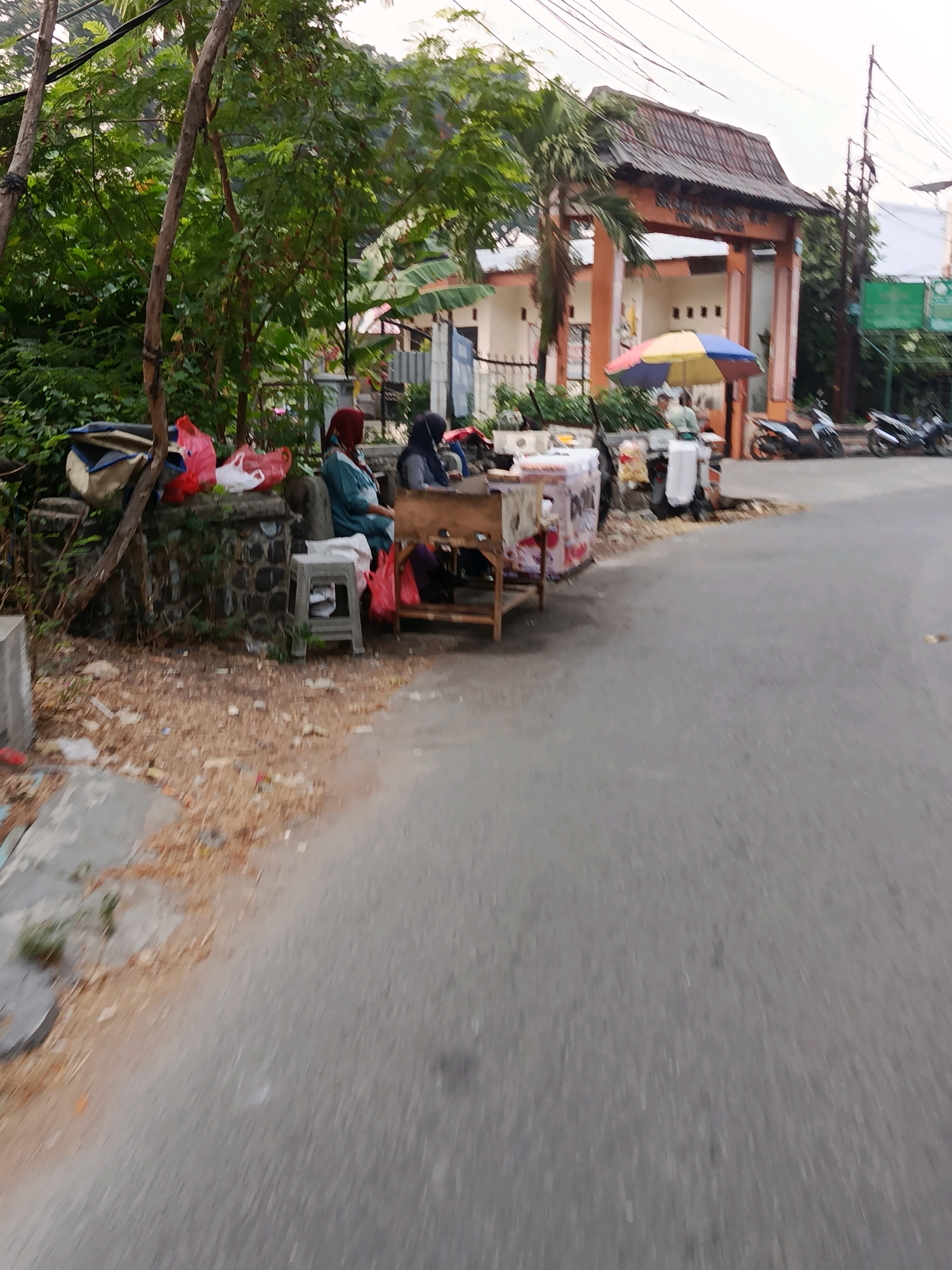 In Bekasi, Indonesia, on a paved road with a slightly cracked surface, a scene unfolds with a focus on a small roadside establishment. A weathered, wooden cart, likely for selling goods, sits prominently. Two women are present. One, wearing a red hijab and a turquoise top, is seated. The other, in a dark hijab and grey top, is also seated, possibly attending to the cart. Red and white plastic bags are visible, some placed on a low, stone wall adorned with moss and debris. A grey plastic stool is positioned near the cart. The background features lush green trees and a building with a distinctive, aged roof structure, possibly an archway entrance to a compound. A colorful, striped umbrella is deployed, providing shade. Several motorcycles are parked on the side of the road, indicating a common mode of transportation. The overall impression is a snapshot of daily life and commerce at the edge of a community.