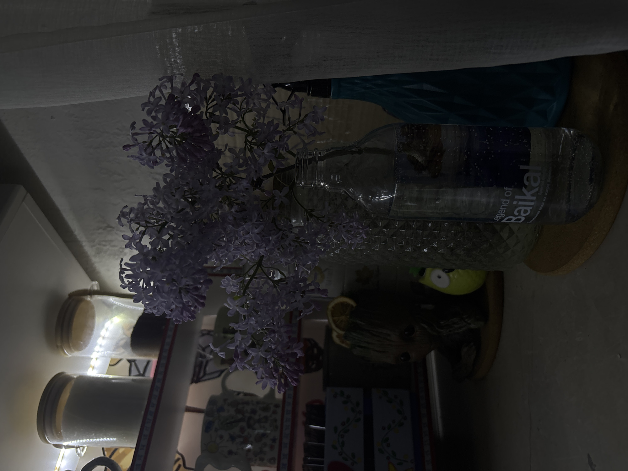 The image, rotated 90 degrees clockwise from its natural orientation, depicts an indoor still life scene. At its center is a fresh bouquet of light purple lilac flowers, arranged in a clear, textured glass bottle. The bottle is a repurposed "Legend of Baikal" mineral water bottle, partially filled with water, and displays the brand name clearly.
To the left of the flowers, two transparent canisters with light-colored, possibly wooden or cork, lids are stacked. They contain a white powdery substance and are adorned with warm-toned LED string lights, which provide localized illumination. Below the canisters, a patterned mug with blue and white designs is partly visible.
Behind the floral arrangement, a blue plastic bag or container can be seen. To the right of the flowers, a small, yellow-green character figurine with large white eyes peeks out, alongside a darker, possibly ceramic, decorative item resembling an animal head topped with a lemon slice. The entire setup rests on a wooden surface. In the upper background, a sheer, light-colored curtain or fabric is visible, possibly near a window.
The scene is dimly lit, suggesting either evening or night, or an interior space with limited natural light, with the LED string lights adding a gentle glow. The overall mood is quiet and domestic. The "Legend of Baikal" branding on the bottle is a specific location cue consistent with Zheleznogorsk, Russia. The visible text on the bottle reads "Legend of Baikal" in English and includes Cyrillic text underneath.