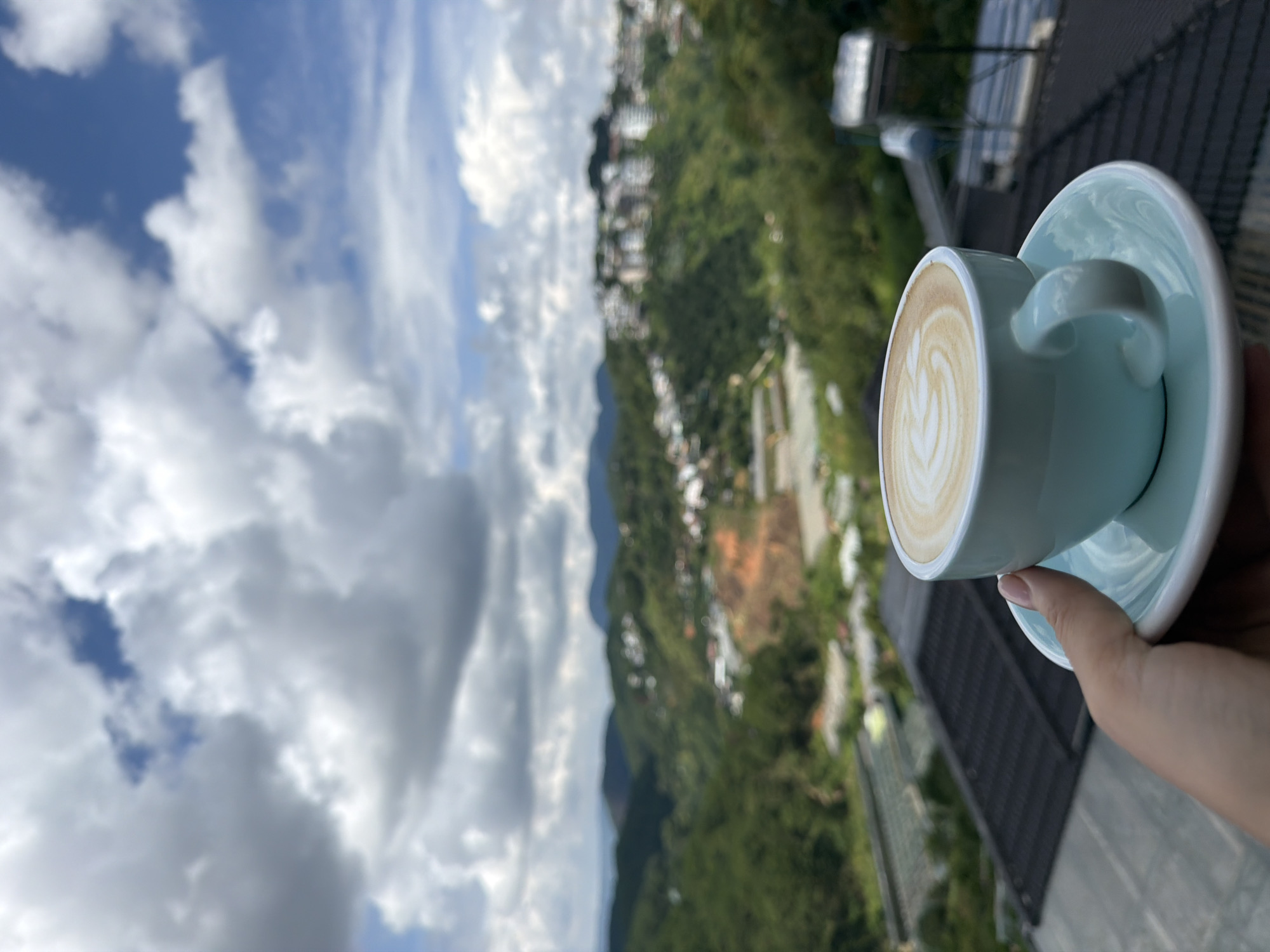 A hand holds a light blue ceramic cup and saucer containing a latte with intricate foam art. The cup is positioned in the foreground, slightly to the right of center. In the background, a sprawling city with numerous buildings is nestled within lush, rolling green hills, indicative of Da Lat, Vietnam. The sky above is a dramatic display of clouds and patches of blue. The viewpoint suggests the photo was taken from an elevated position, possibly a balcony or cafe overlooking the city and its surrounding landscape.