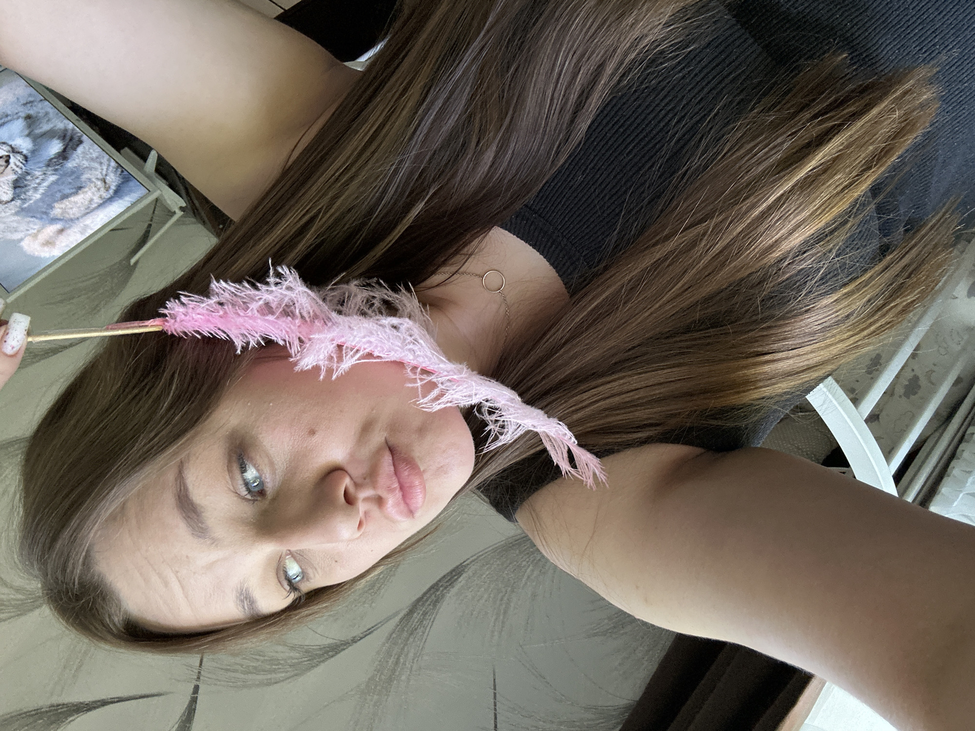 A woman with long brown hair is holding a pink, feather-like object near her face. She has fair skin and light-colored eyes. She is wearing a black top and a delicate necklace with a circular pendant is visible. A digital tablet is propped up in the background. The location is likely indoors in Chaikovsky, Russia.
