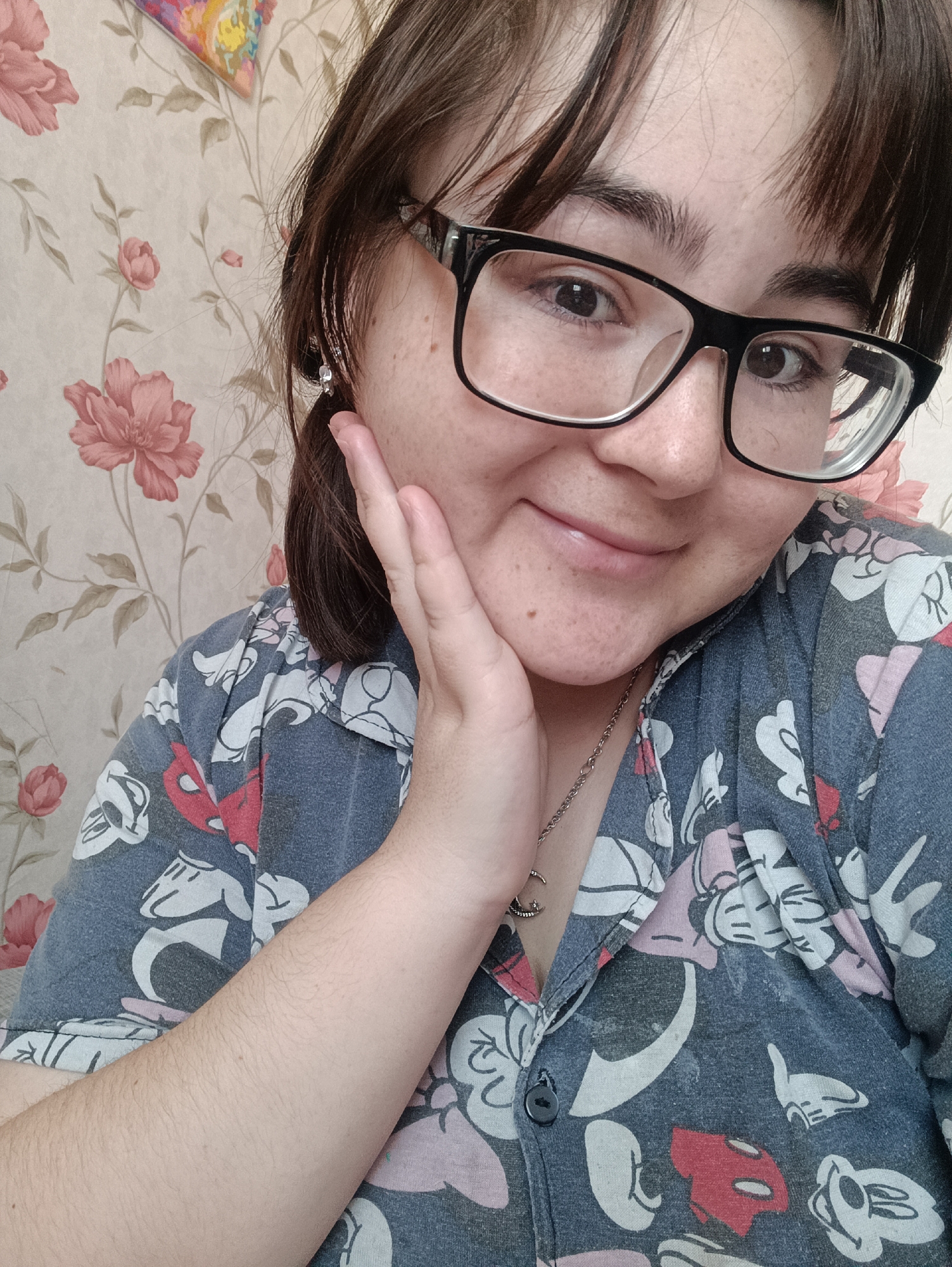 A close-up of a woman wearing glasses and a button-down shirt with cartoon characters is shown indoors. The background is patterned wallpaper with pink floral designs. The woman is touching her face with her right hand. She has dark hair and light skin. A silver necklace is visible around her neck. The specific location is not discernible from the image.