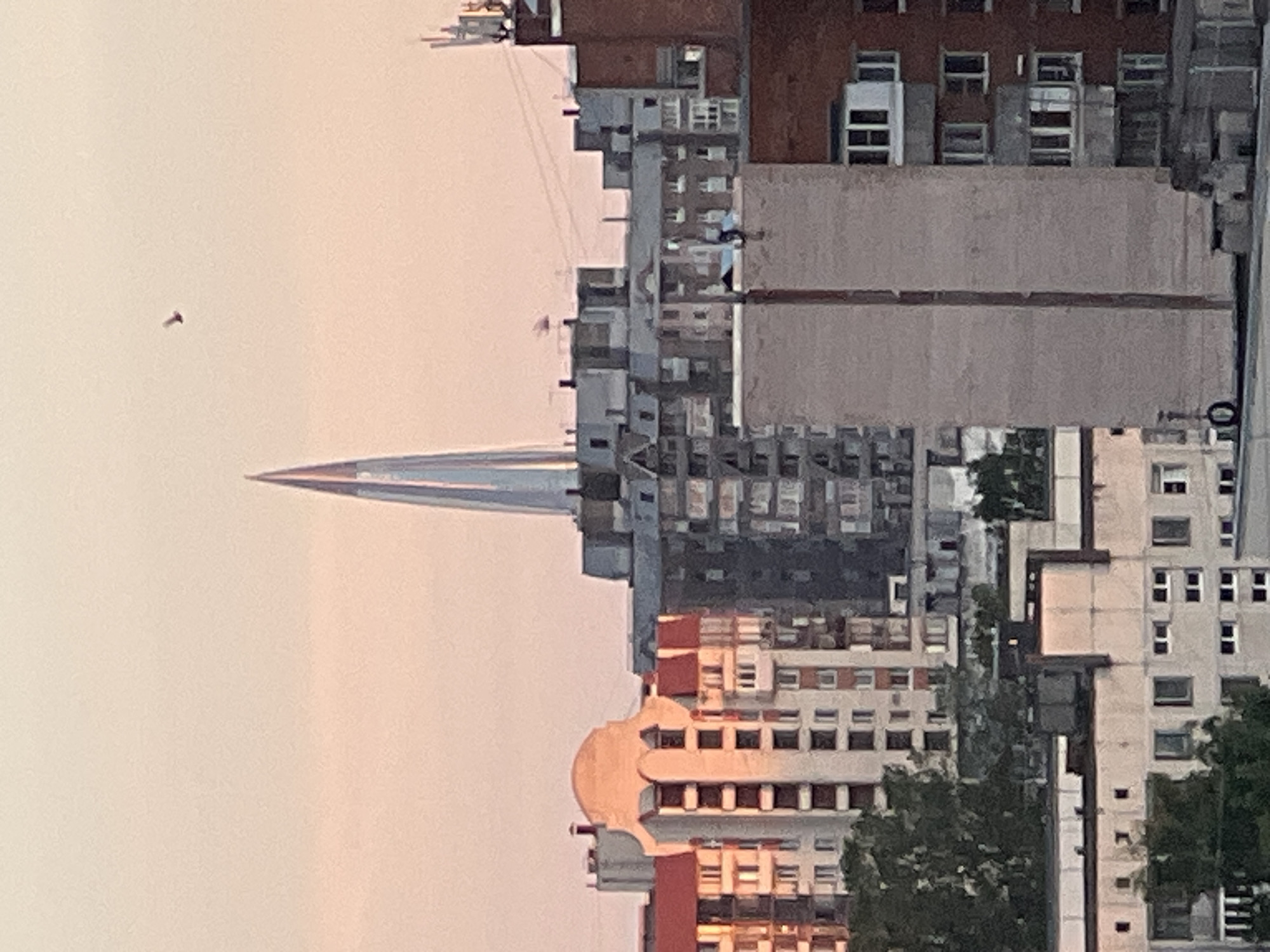 This is a vertical, slightly blurry, long-shot photograph of a city skyline at dusk. The image is oriented such that the buildings are upside down. Dominating the left side of the frame is a tall, needle-like structure that tapers to a sharp point, reflecting the soft, pinkish glow of the sunset. Surrounding this central spire are numerous multi-story buildings, characterized by repetitive window patterns and a mix of architectural styles, suggesting an urban environment. The buildings appear to be made of concrete and brick, with some having lighter-colored facades.
In the upper left portion of the image, a small, dark object, possibly a bird or a drone, is visible against the pale sky. On the right side of the frame, taller buildings with more intricate details are present, including balconies and various window designs. Some trees with dark green foliage are interspersed between the buildings, hinting at parks or street greenery. The sky is a gradient of soft pinks and oranges, indicative of either sunrise or sunset. There is no visible text in the image. The overall impression is one of a dense urban landscape viewed from a distance during a calm, transitional time of day.