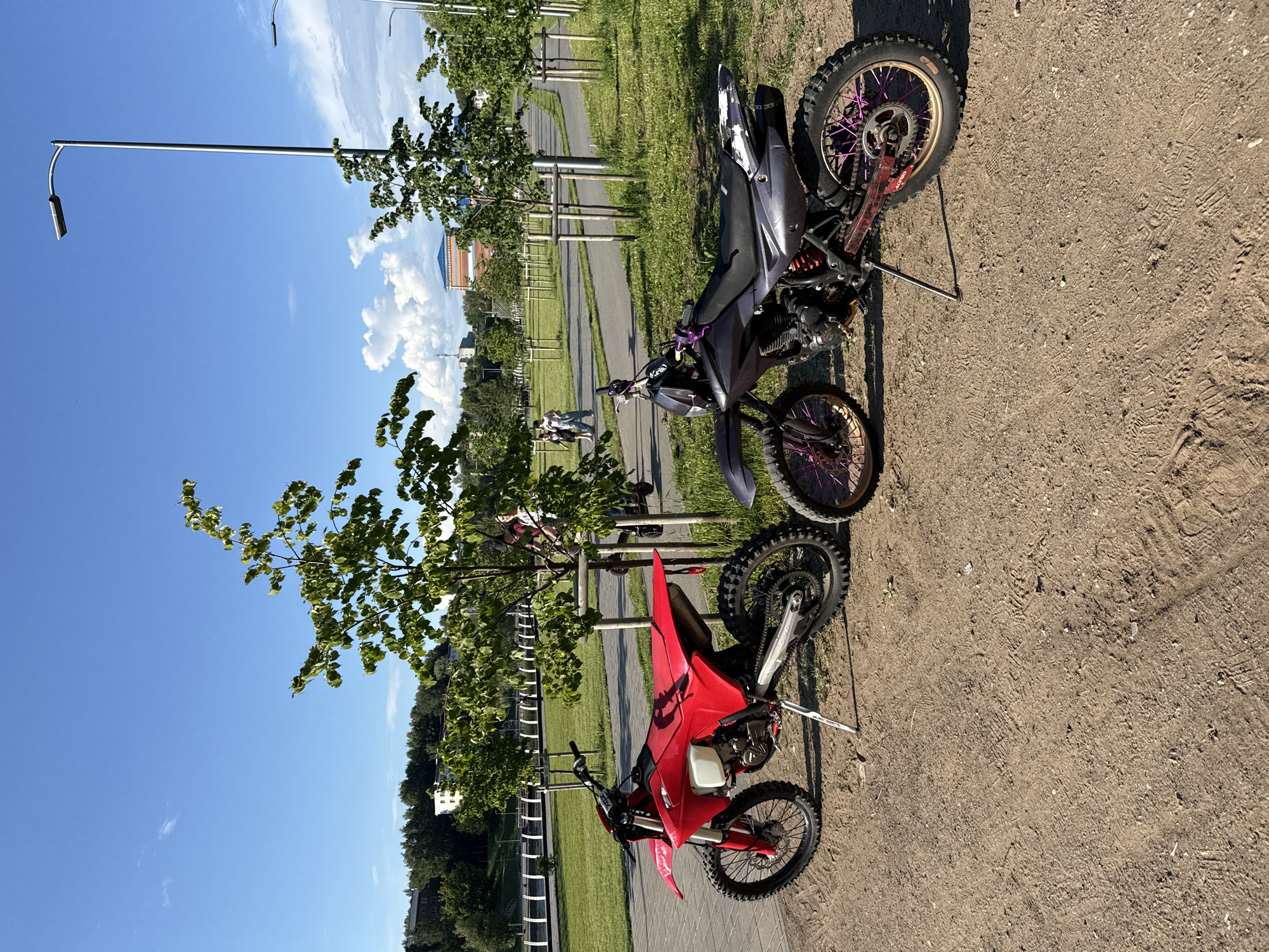 Two dirt bikes are parked on a dirt path in a park on a sunny day. The bike on the left is red and parked on its kickstand. The bike on the right is dark gray with purple accents and is also parked on its kickstand. In the background, a paved path winds through a grassy area with trees. Several streetlights are visible along the path. In the far background, buildings and a radio tower can be seen. A few people are walking in the background, some on scooters. The scene appears to be in the city of Votkinsk, Russia, likely in a park or recreational area near a body of water, given the distant embankment and fence.