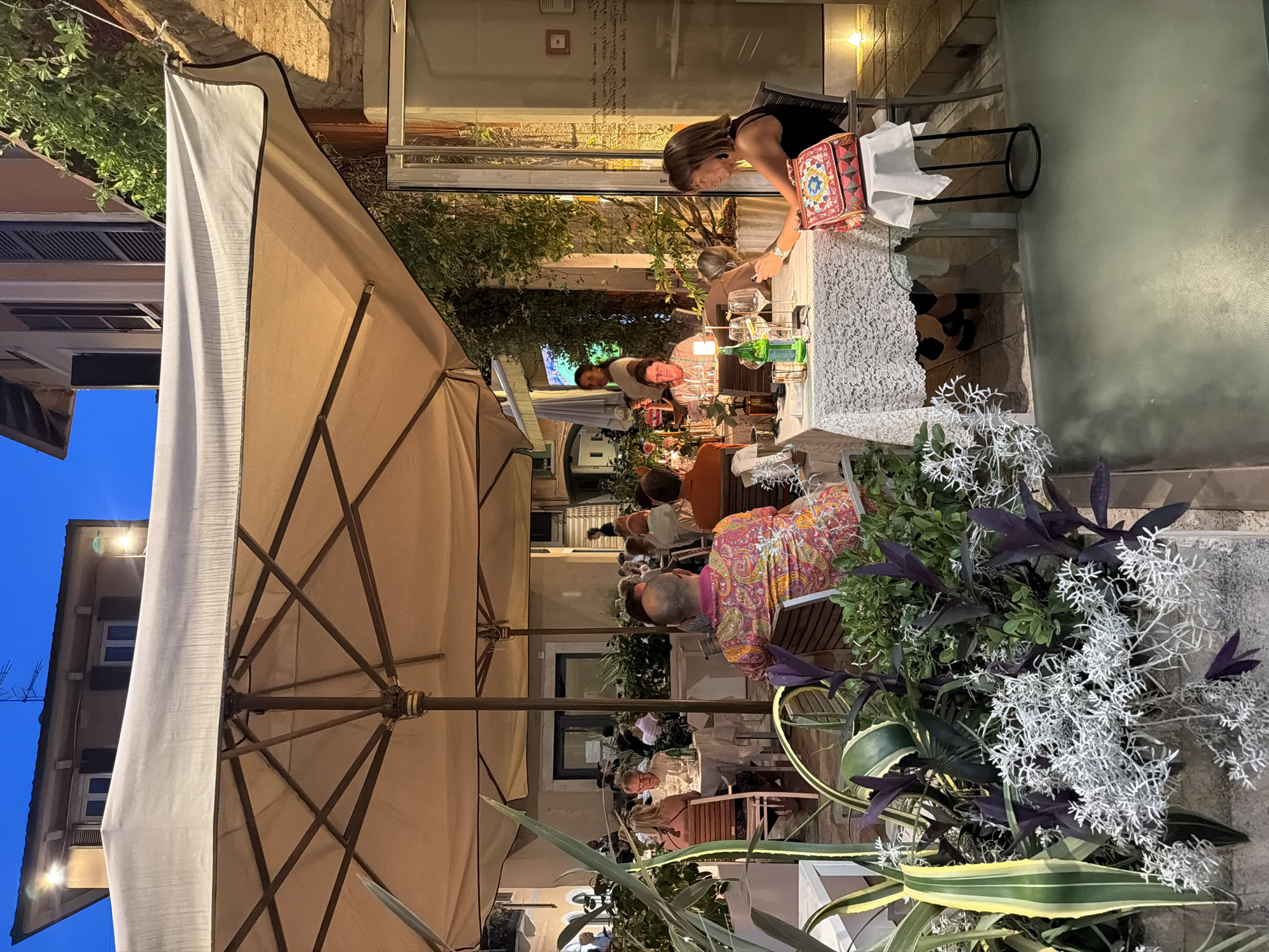 The photograph captures an outdoor dining scene at dusk in Salò, Italy. A large, beige umbrella shades several tables occupied by diners. In the foreground, a woman with shoulder-length brown hair, wearing a black sleeveless top, sits at a table covered with a white lace tablecloth. She is looking at her phone and has a colorful, patterned handbag placed on a small white stand next to her. On the table are wine glasses, a bottle of green liquid, and other dining accouterments. Behind her, other patrons are seated at tables under the umbrella, some engaged in conversation. Lush green foliage is visible surrounding the dining area, and a building with windows and shutters stands in the background. The lighting suggests it is early evening, with artificial lights illuminating the tables and the building.