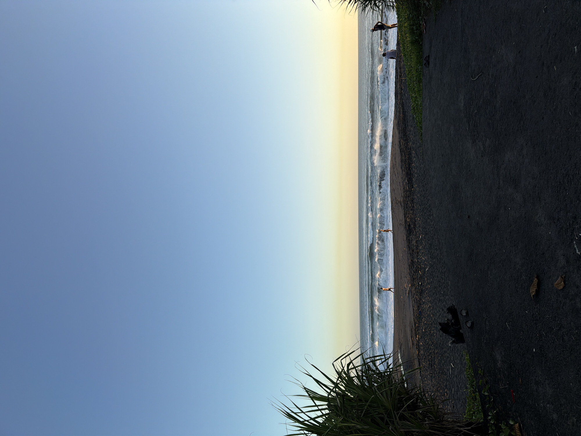 The photograph captures a beach scene near Kediri, Indonesia. A wide expanse of the sky, transitioning from light blue to yellow, dominates the left side of the frame. The beach itself runs vertically through the center, featuring dark volcanic sand and ocean waves. Several individuals are visible wading in the water. Adjacent to the beach is a dark pathway bordered by vegetation. Palm-like plants appear in the foreground and along the path.