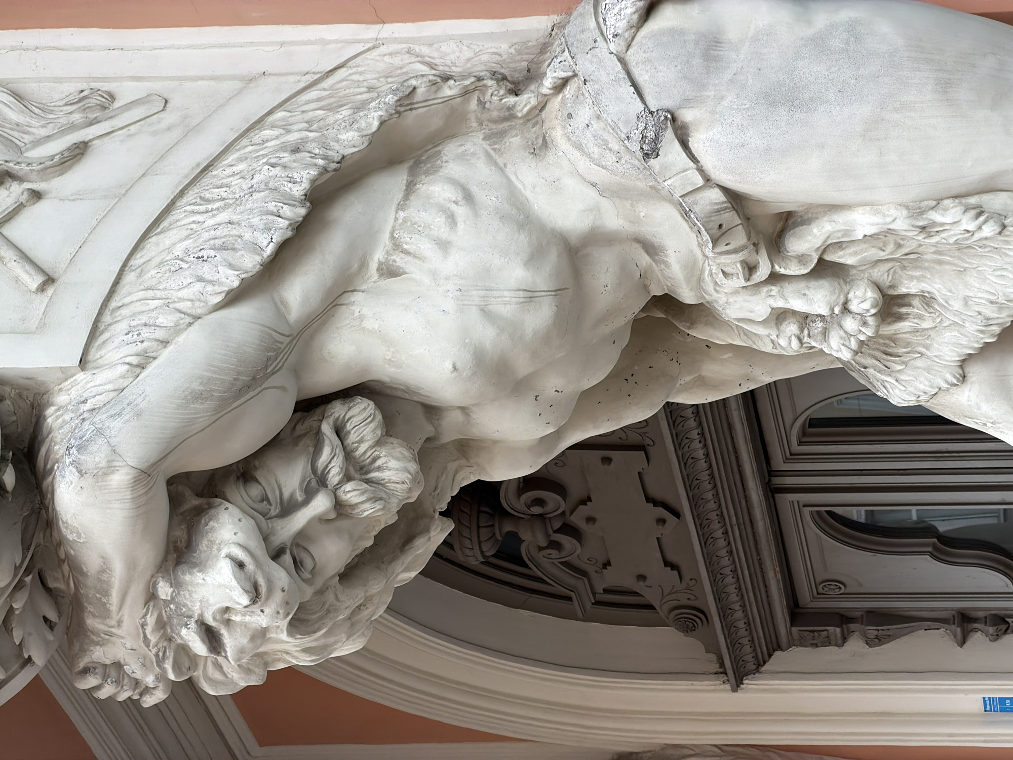 This close-up photograph captures a sculptural element, specifically a telamon, in Saint Petersburg, Russia. The sculpture depicts a muscular, nude male figure with a lion's head covering part of his head and shoulders. A belt or harness appears to be securing the lion pelt to the man's body. The figure is positioned horizontally, appearing to support a structure above. The telamon is made of a light-colored material, possibly stone or plaster, with visible weathering and some surface discoloration. Architectural details are also visible in the background, indicating it is part of a building facade. These details include decorative moldings and the corner of a window with a light gray frame. A faded salmon-colored surface is evident behind the sculpture, likely the exterior wall of the building. A blue sticker is visible towards the bottom right corner of the image.