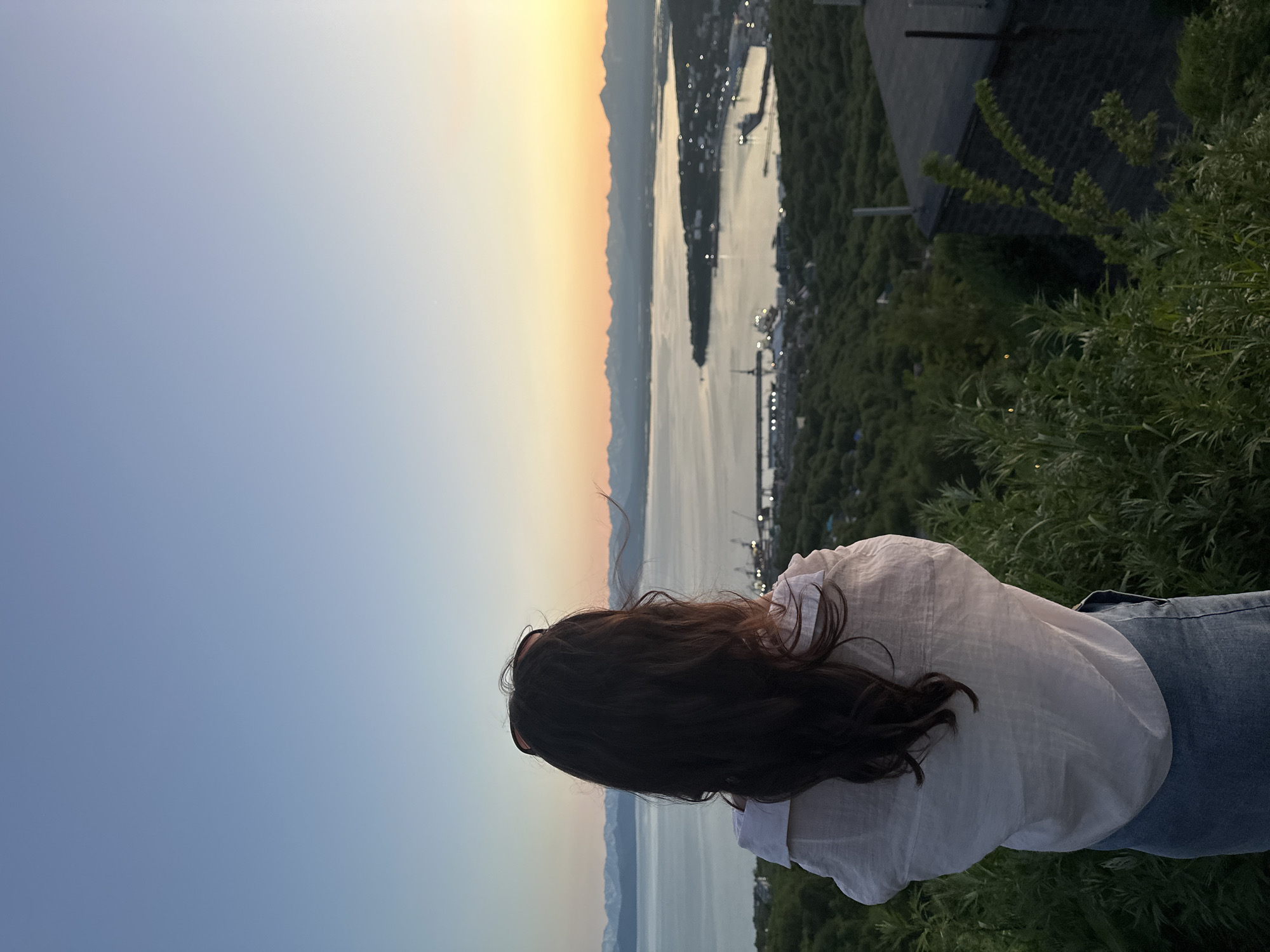 A young woman with long, dark hair stands with her back to the camera, looking out over a bay at sunset. She is wearing a loose-fitting white shirt and jeans. In the distance, a city is nestled along the coastline, with lights twinkling in the twilight. Beyond the city, a body of water stretches out to meet a range of snow-capped mountains on the horizon. The sky above transitions from a pale blue near the top of the frame to shades of orange and pink near the horizon, indicating the sun has recently set or is about to set. The foreground is dominated by lush green foliage and the corner of a dark-colored building, suggesting the viewpoint is from a hilltop overlooking the city of Petropavlovsk-Kamchatsky, Russia.