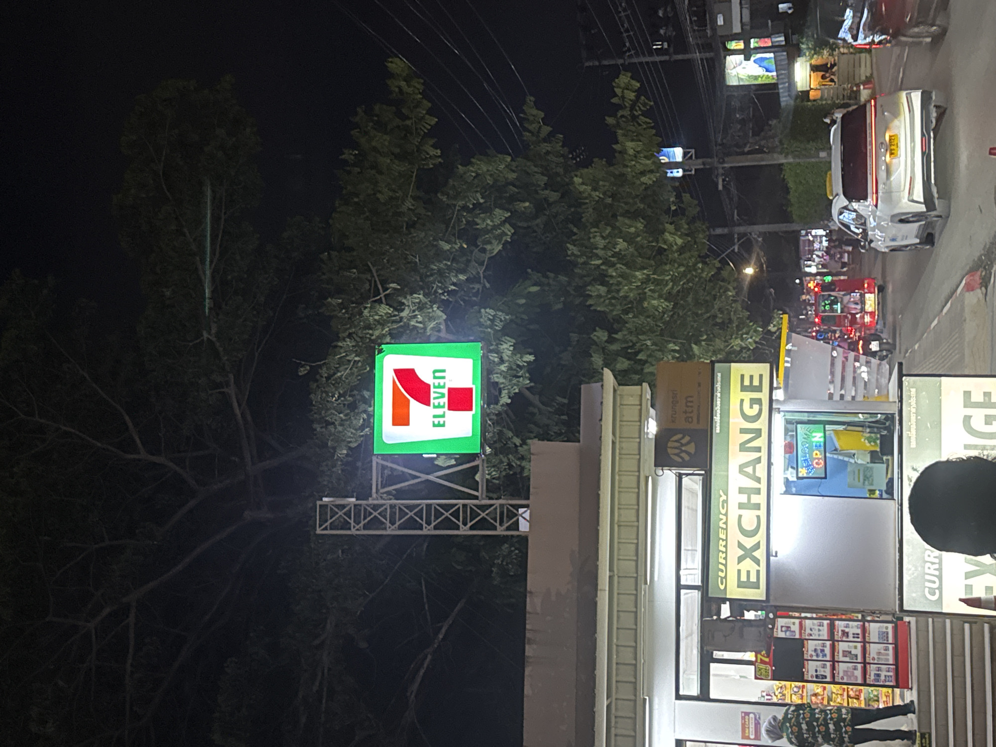 Here is a description of the image:
The image shows an intersection in Patong, Thailand, at night. There is a 7-Eleven convenience store on the left, with its illuminated green sign visible above a Krungsri ATM and currency exchange booth. People are shopping in the 7-Eleven and standing near the Currency Exchange booth. Traffic is visible in the distance, including cars and scooter taxis. Trees are on the left of the frame behind the 7-Eleven sign. Powerlines cross the sky. Streetlights illuminate the scene.