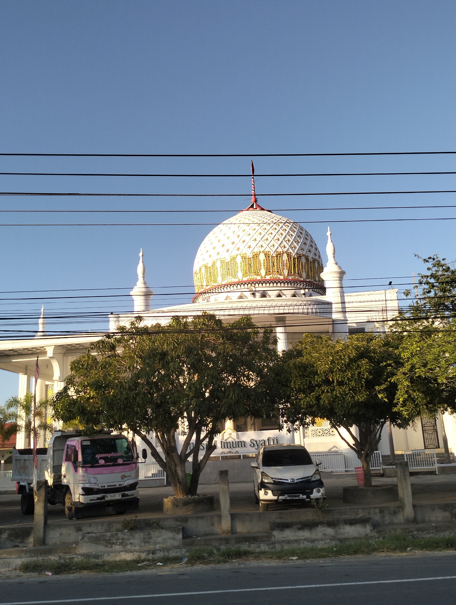 The image depicts the exterior of a white mosque with a large, ornate dome, topped with a red finial. The dome is decorated with intricate golden patterns and white diamond shapes. Two smaller white minarets flank the main dome. The mosque features a white facade with arched openings and decorative latticework. In front of the mosque, several trees with dense green foliage are visible, casting shadows on the ground.
To the left, a white and pink dump truck is parked. The truck has decals on its windshield and side. To the right of the truck, a silver minivan is parked under a tree. Both vehicles are facing towards the viewer.
The scene is set against a clear blue sky. Several black power lines traverse the upper portion of the image, obscuring parts of the sky and the mosque's dome and minarets. A low concrete barrier runs along the foreground, separating the road from the area where the vehicles and mosque are situated. The ground in front of the mosque appears to be paved or covered in gravel.
The image is a photograph taken outdoors, likely during daylight, given the clear sky and the presence of shadows. The overall impression is one of a religious building and its surroundings in a developed area.