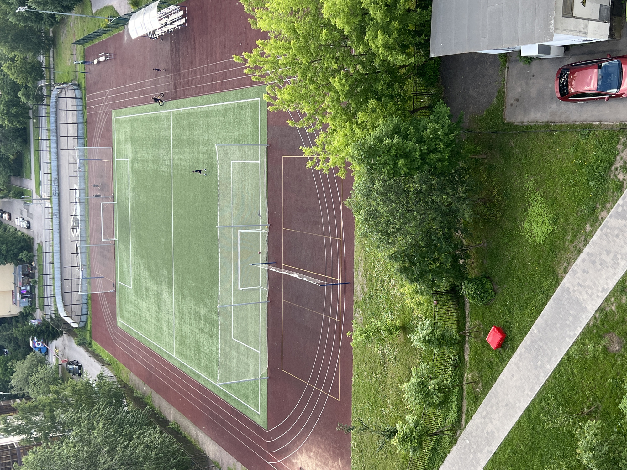 An aerial view captures a sports complex located in a city in Russia. The complex features a green artificial turf soccer field with white boundary lines and two soccer goals. Adjacent to the soccer field is a red running track with four lanes, which curves around the field. A net divides a section of the running track into what appears to be a volleyball court. In the background, a fenced-off area contains a dirt surface, possibly a basketball court, and a seating area with bleachers. Several people are present on the grounds, including individuals walking on the track, one riding a bicycle, and a few standing near the soccer goals. Surrounding the sports complex are residential buildings and trees. A red car is parked near a building on the lower right side of the image, and a red cube-shaped object rests on a grassy area to the left of a paved walkway.