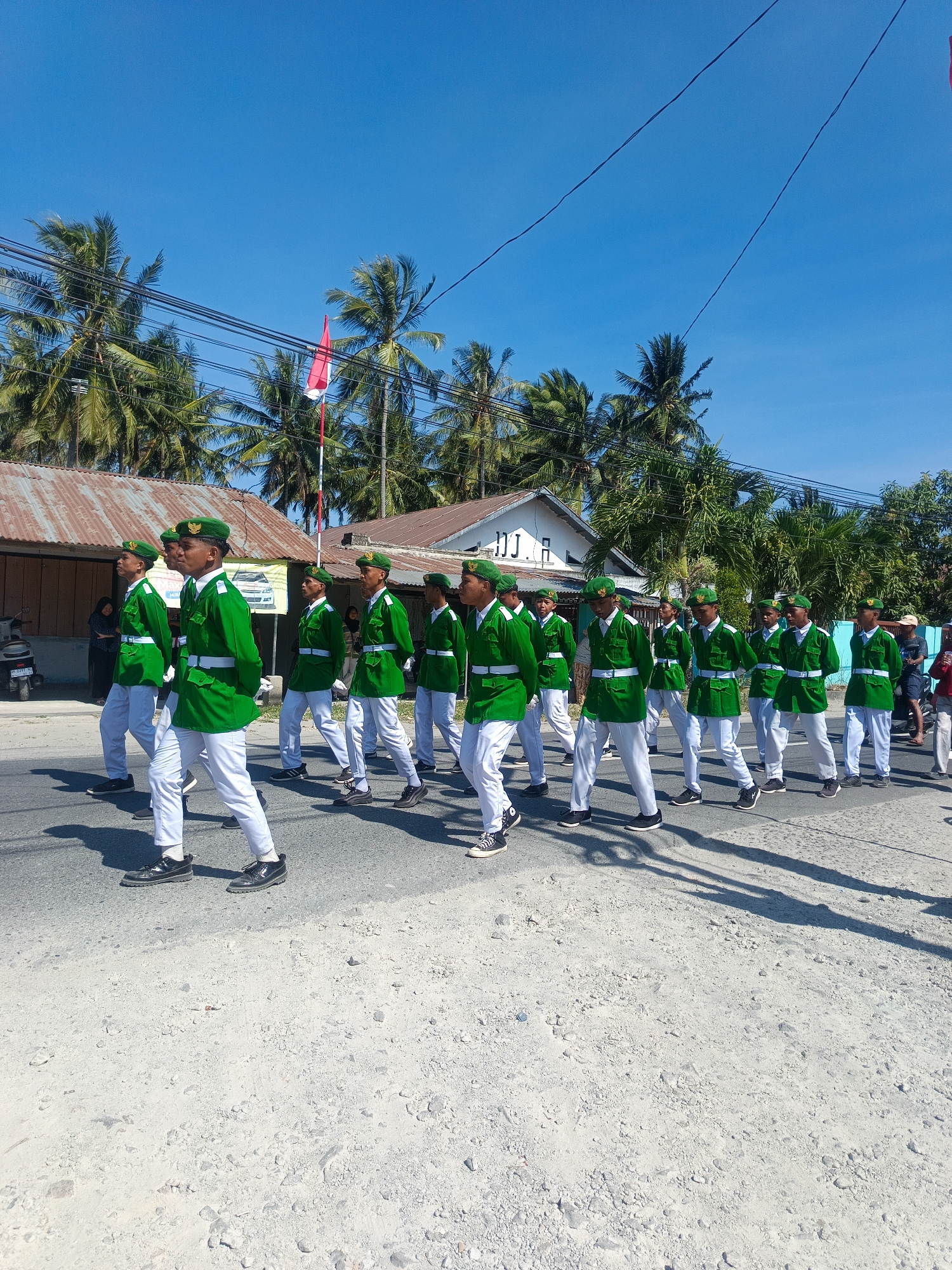 In Tenggela, Indonesia, a group of young males, dressed in matching green jackets, white pants, and green berets, are marching in formation on a paved road. They are wearing white belts over their jackets. The individuals are of varying heights and appear to be in good health. Some are looking forward, while others are looking to the side or down. The group is marching in an organized manner, with several rows of individuals. The environment is outdoors, with a clear blue sky overhead. Coconut trees are visible in the background, along with some buildings. A red and white flag is also visible in the background, flying from a pole. The sun is shining brightly, casting shadows on the road. A motorcycle is parked on the left side of the image. The road surface appears to be asphalt, with some gravel and sand on the sides. The overall scene suggests a parade or a formal procession.