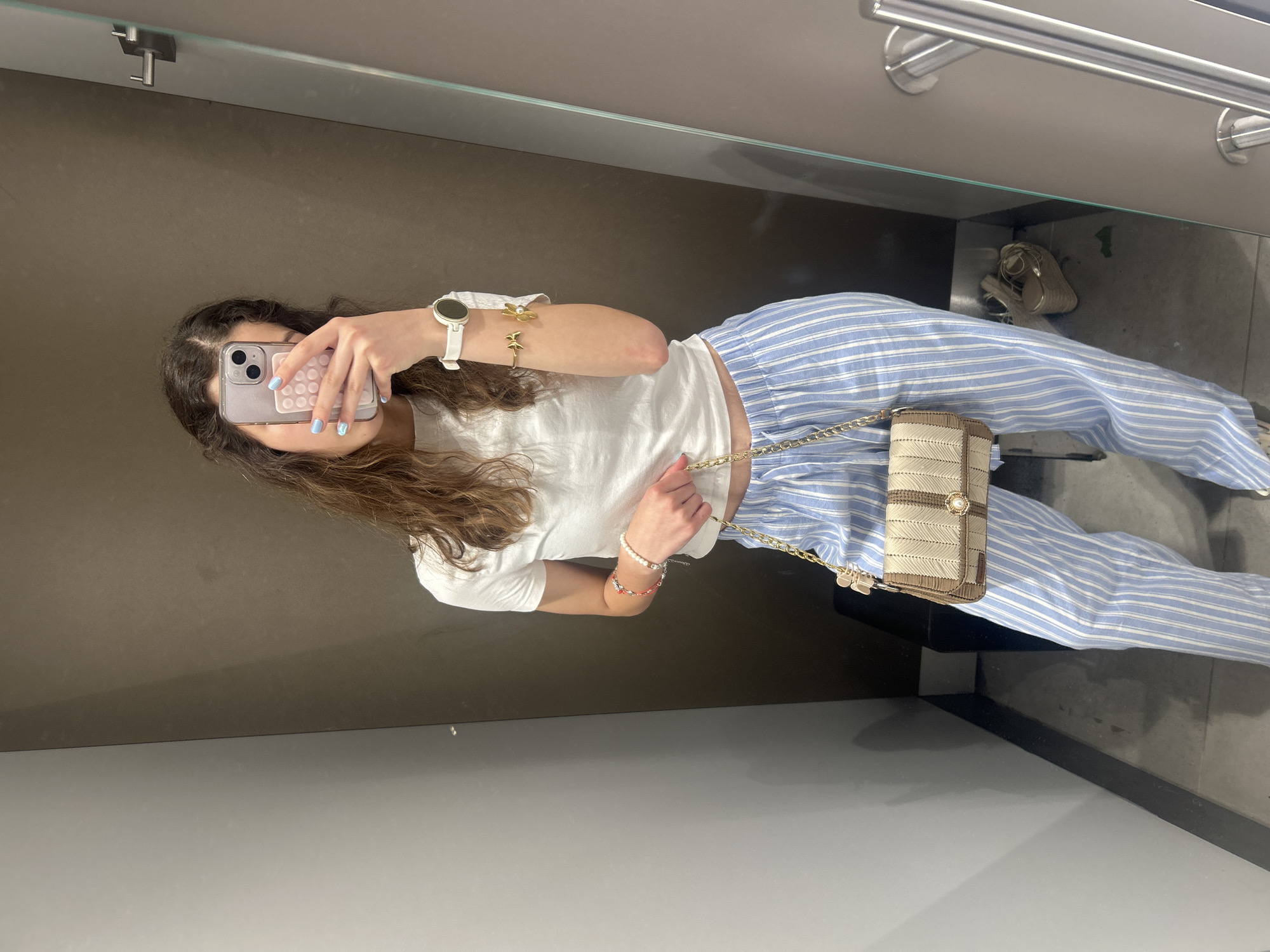 A young woman with long, curly brown hair is taking a mirror selfie in what appears to be a fitting room. She is wearing a white t-shirt, light blue and white striped pants, and a small beige purse with a gold chain. She is also wearing a white smartwatch on her left wrist and several bracelets. Her fingernails are painted light blue. In the background, a mirror reflects her image and part of the room, which has grey and brown walls and a metal clothing rack. There are also a pair of gold wedge sandals visible on the floor. The woman is holding her phone with her right hand, which is angled to capture the selfie. The scene does not provide any specific visual cues that can be used to determine a location in the city of Ruse, Bulgaria.