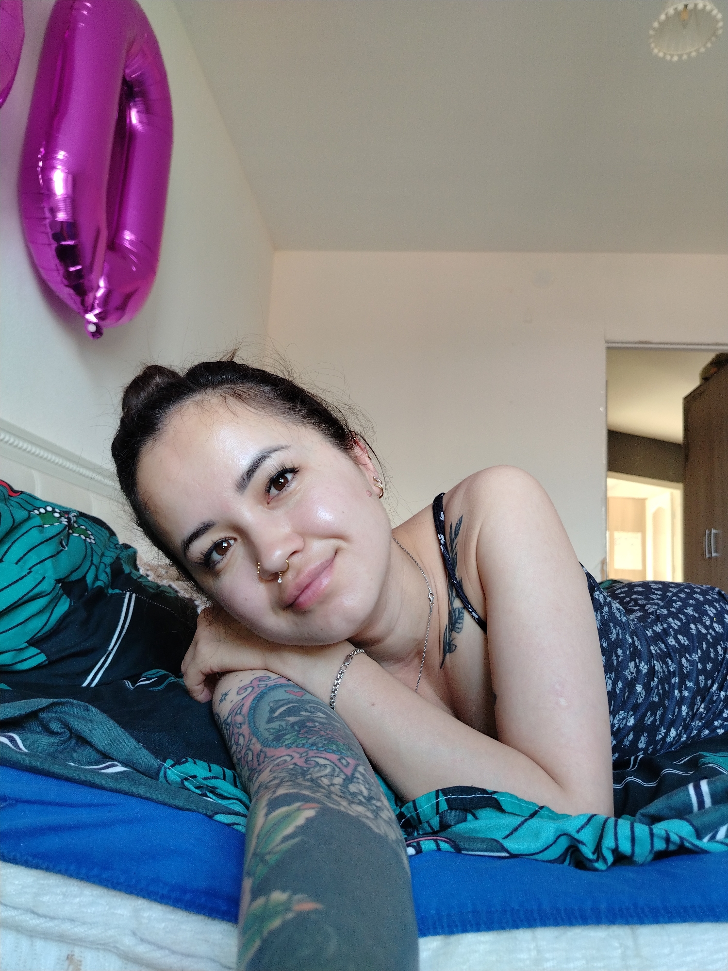 A young woman with dark hair pulled into a bun lies on a bed. She is wearing a dark floral print dress. Her left arm, covered in tattoos, is visible, and her head is propped on her right arm, which is bent at the elbow. She has a nose ring and earrings. Two large, shiny magenta balloons shaped like the number "0" are attached to a white wall in the background. The room appears to be a bedroom with bedding and a portion of a closet visible in the background. The image is taken from a slightly high angle, looking down at the woman. It is not possible to determine the specific location in the city of Noviy, Russia, from the image alone.