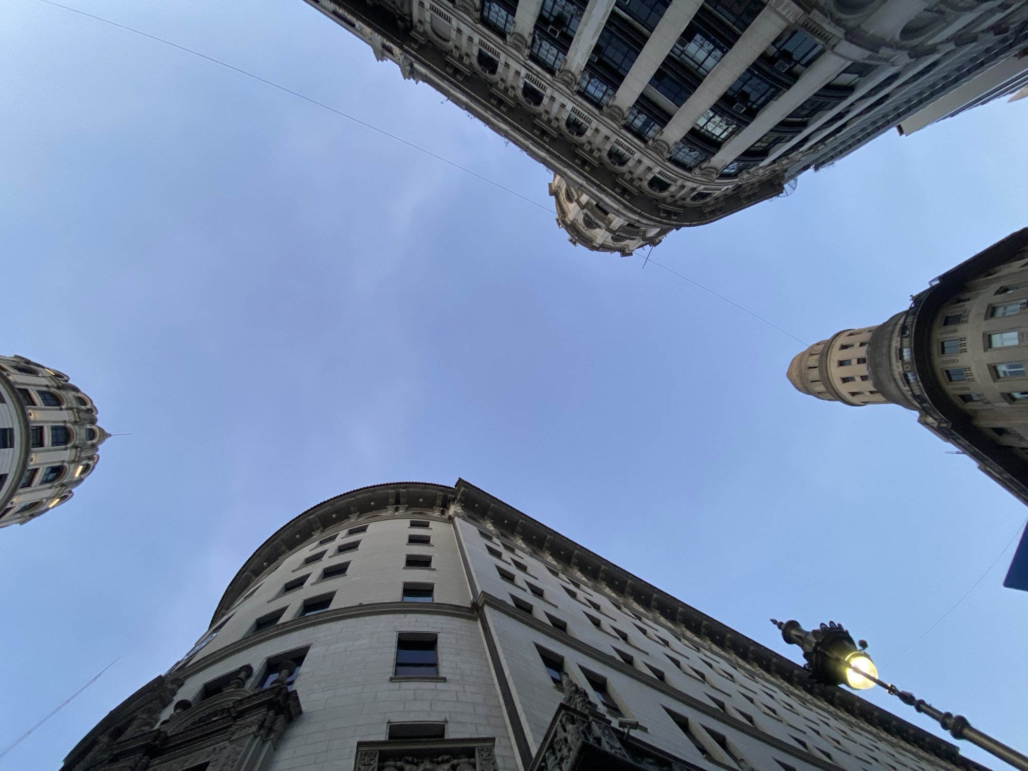 The image is a low-angle shot of four buildings in Buenos Aires, Argentina. The buildings are architectural landmarks with ornate facades. The sky is visible in the spaces between the buildings and is a pale blue color. Wires are strung between the buildings. A street lamp is visible in the lower right corner of the frame. There are no people visible in the image.