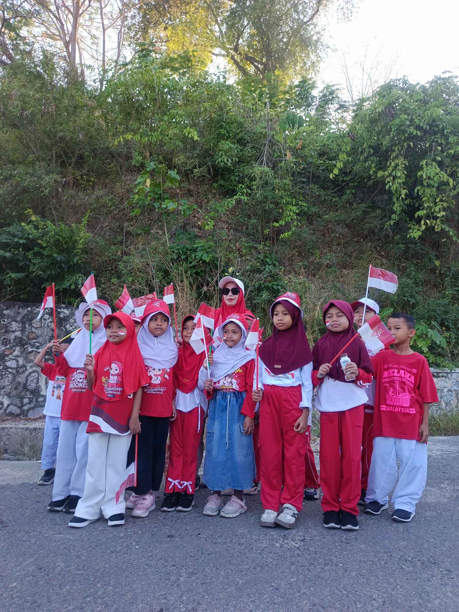 In Ilotidea, Indonesia, a group of approximately ten children, mostly girls, and one adult woman stand in the foreground on a paved surface, likely a road. The children are dressed in red and white, many wearing red shirts and white or red pants. Several children are wearing hijabs, also in red or white. They are holding small Indonesian flags, with red and white patterns, on sticks. One child has face paint on their cheeks, resembling the Indonesian flag. The adult woman, at the far right, is wearing a red hijab and sunglasses.
Behind the group, a steep, overgrown hillside is visible, covered in dense green foliage, bushes, and trees. The lighting suggests it is daytime, possibly late afternoon, with sunlight filtering through the leaves of the trees at the top of the hill. The overall scene appears to be a patriotic gathering, possibly related to a national holiday or celebration, given the prominent display of Indonesian flags and the national colors. The environment is natural and somewhat rugged, with the paved road contrasting with the wild vegetation.