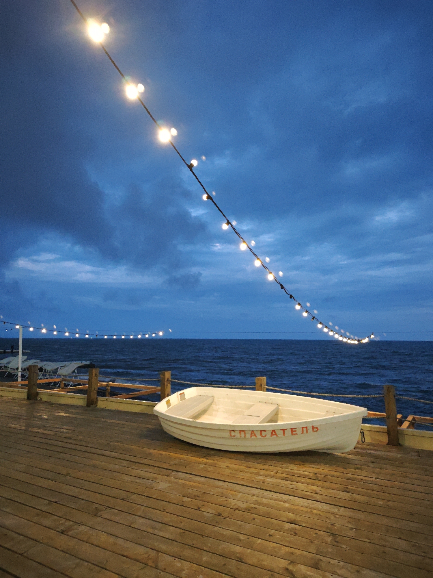 The image captures an evening scene on a wooden pier in Sochi, Russia. A white lifeguard boat with the word "СПАСАТЕЛЬ" (Rescuer) in red Cyrillic letters is docked on the pier. Above, a string of lights illuminates the scene, with the dark sea and cloudy sky in the background. In the distance, another line of lights stretches along the coast. There are no people visible in the immediate foreground, but chairs can be seen in the distance.