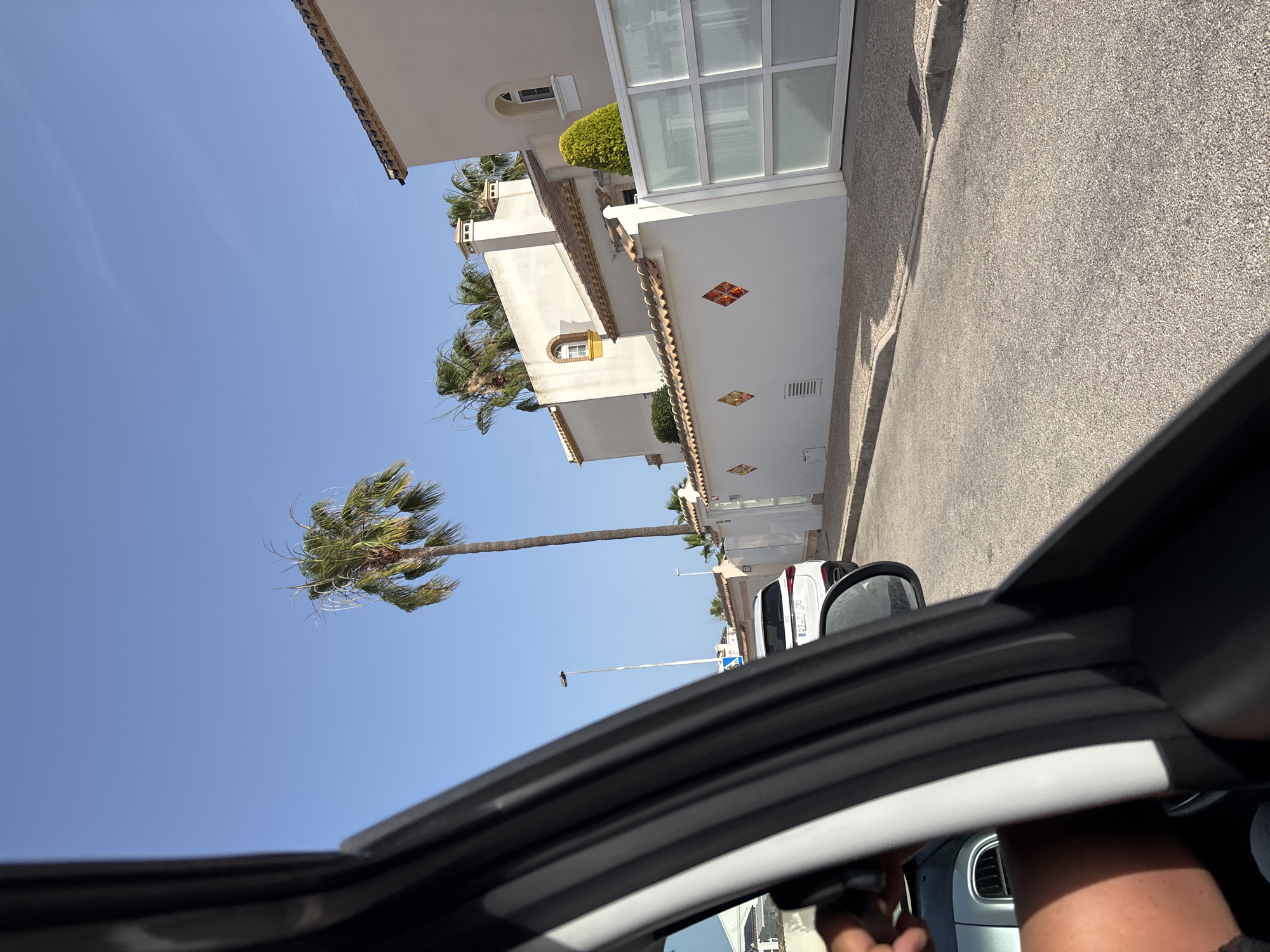 The image shows a view from inside a vehicle looking out towards a street lined with modern houses and palm trees. The houses are white with terracotta tiled roofs and feature decorative diamond-shaped tiles on the front wall. A white SUV is parked on the street. The sky is clear blue. The scene appears to be a residential area, likely in a warm climate. The location is identified as Orihuela, Spain.