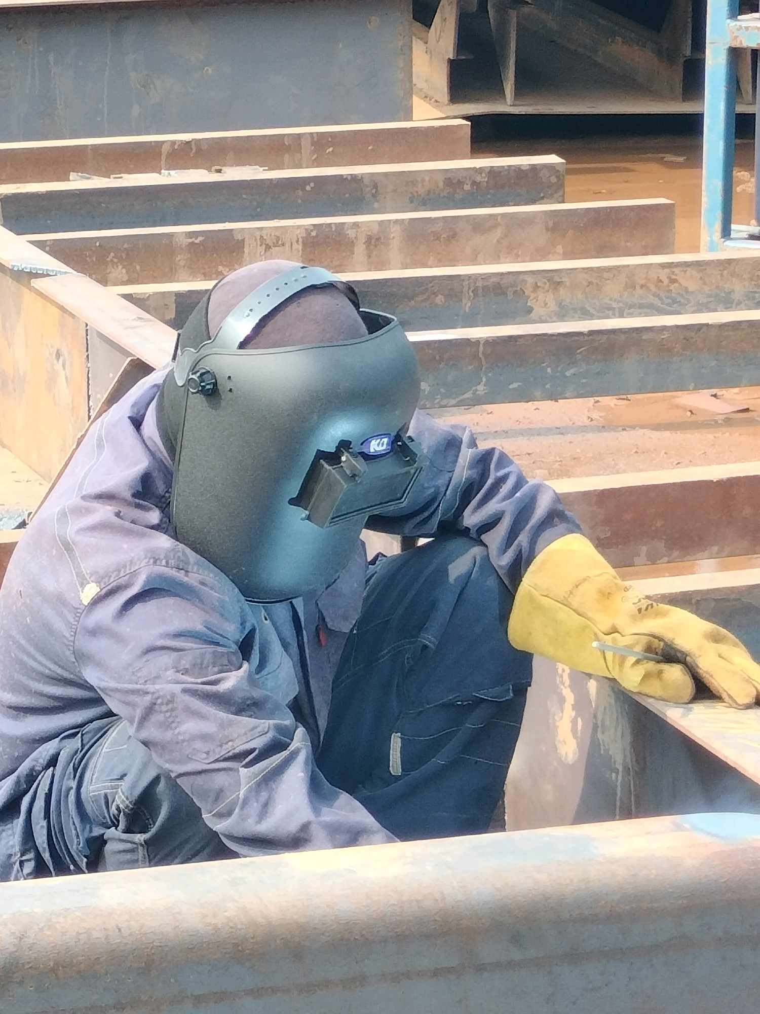 A person wearing a welding mask and a blue uniform, complete with yellow gloves, is working on a metal structure in Batam, Indonesia. The individual is kneeling and appears to be holding a welding rod. The environment suggests a construction or industrial setting with large metal beams and a dusty, possibly metallic, floor. The scene is outdoors under natural lighting.