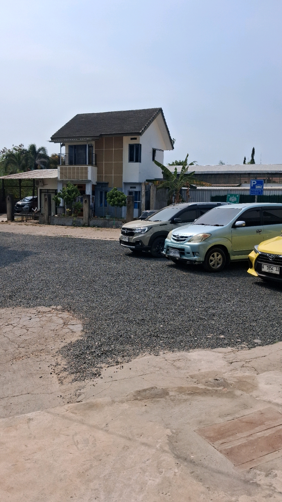 In a parking lot located in Banjarbaru, Indonesia, a two-story house with a white facade and a dark tiled roof stands under a clear sky. The house features a balcony with a metal railing on the second floor. In front of the house, several vehicles are parked on a gravel surface. These include a light-colored SUV, a light blue minivan, and a yellow car, all facing towards the right side of the frame. A parking sign with a blue "P" symbol and the words "PARKIR RODA 4" is visible in the background, near a low building with a corrugated metal roof. The ground in the foreground is a mix of cracked concrete and gravel. Lush green trees and foliage surround the property.