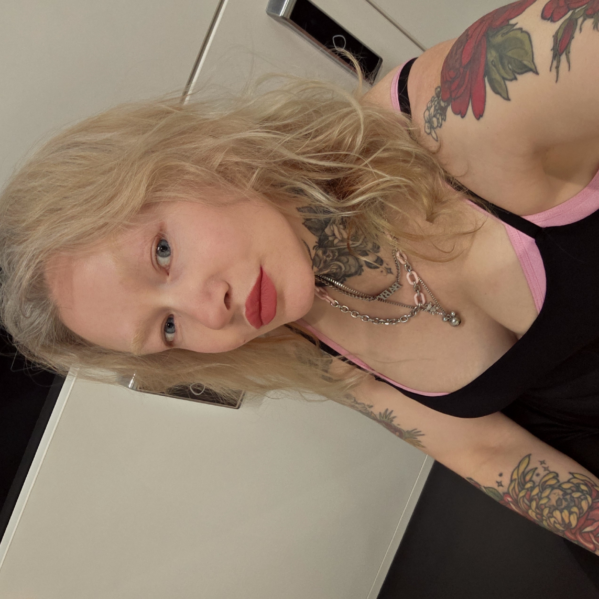 The image presents a close-up portrait of a person, likely female, with long, wavy blonde hair and light blue eyes. She has fair skin and is wearing red lipstick, with a neutral to slightly contemplative expression as she looks directly at the camera.
The individual is heavily tattooed. On her right shoulder and upper arm, a large floral tattoo, primarily in red and green hues, is visible. Her left forearm displays another vibrant, colorful floral design featuring yellow and red elements with small star accents. Additionally, intricate black and grey floral or ornate tattoos are visible on her neck.
She is wearing a black top with pink trim around the neckline and straps. Accessorizing her look is a multi-layered silver-toned necklace. One strand features a small bell-shaped charm, while another prominent strand spells out the word "BABY" in block letters.
The background suggests an indoor setting, characterized by light-colored, smooth surfaces, possibly walls or furniture like a cabinet or door. A dark, rectangular object with a metallic handle or accent is partially visible in the upper background. No specific time of day or weather can be discerned from the image. While the image itself provides no location cues, the provided context indicates the scene is located in Saint Petersburg, Russia.