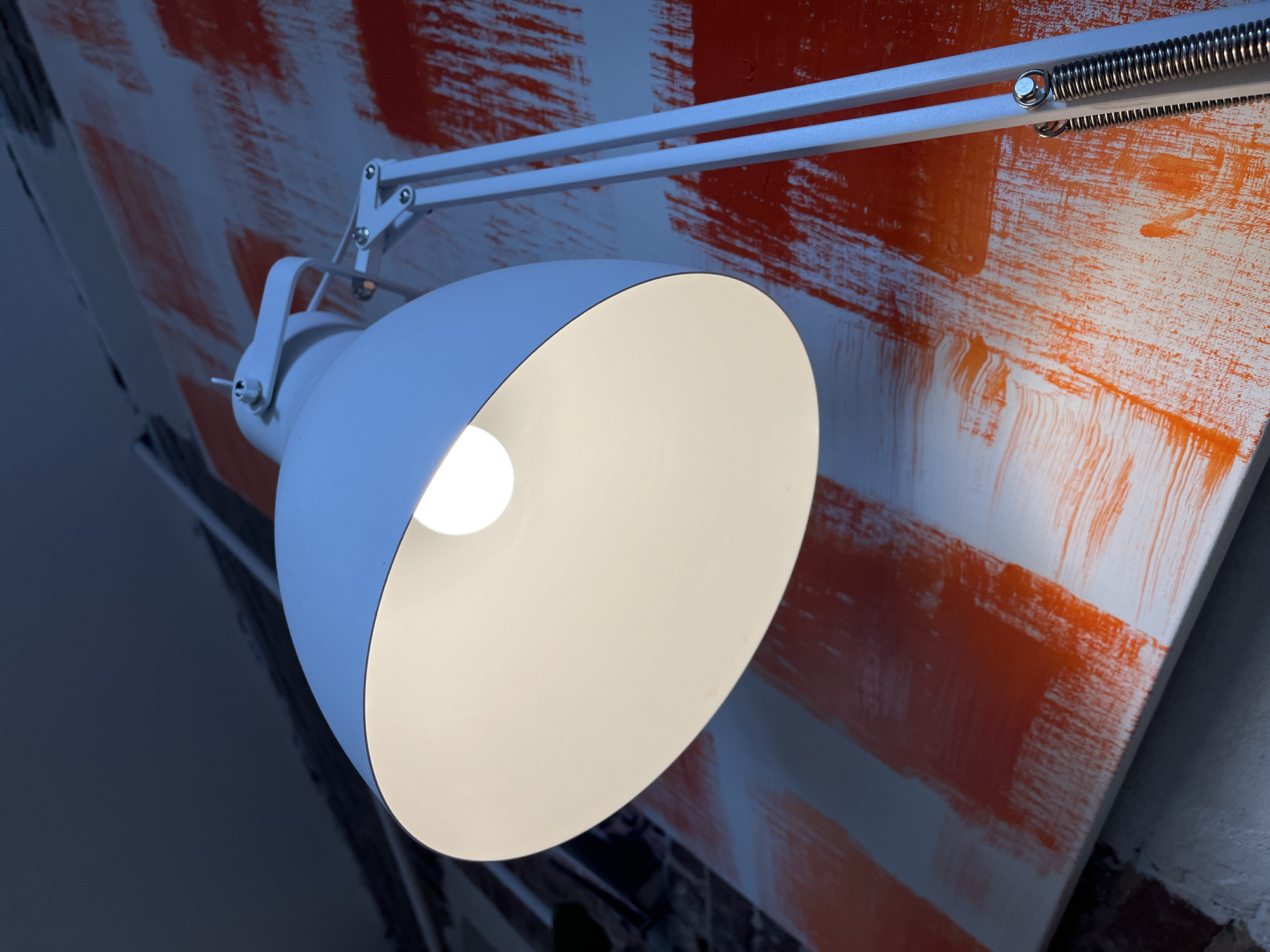 An eye-level, close-up, low-angle shot shows a white desk lamp with its light on, illuminating a canvas in the background. The canvas has an abstract pattern of wide, orange brushstrokes on a white background, arranged in horizontal bands. The lamp has a flexible arm and a wide shade, casting a bright, warm light. The background is dark and out of focus, with hints of brickwork and a ceiling. There are no people or animals visible in the image. The scene is indoors, and the lighting suggests it is likely evening or night. The specific location in the city of Vitsebsk, Belarus, cannot be determined from the image alone.