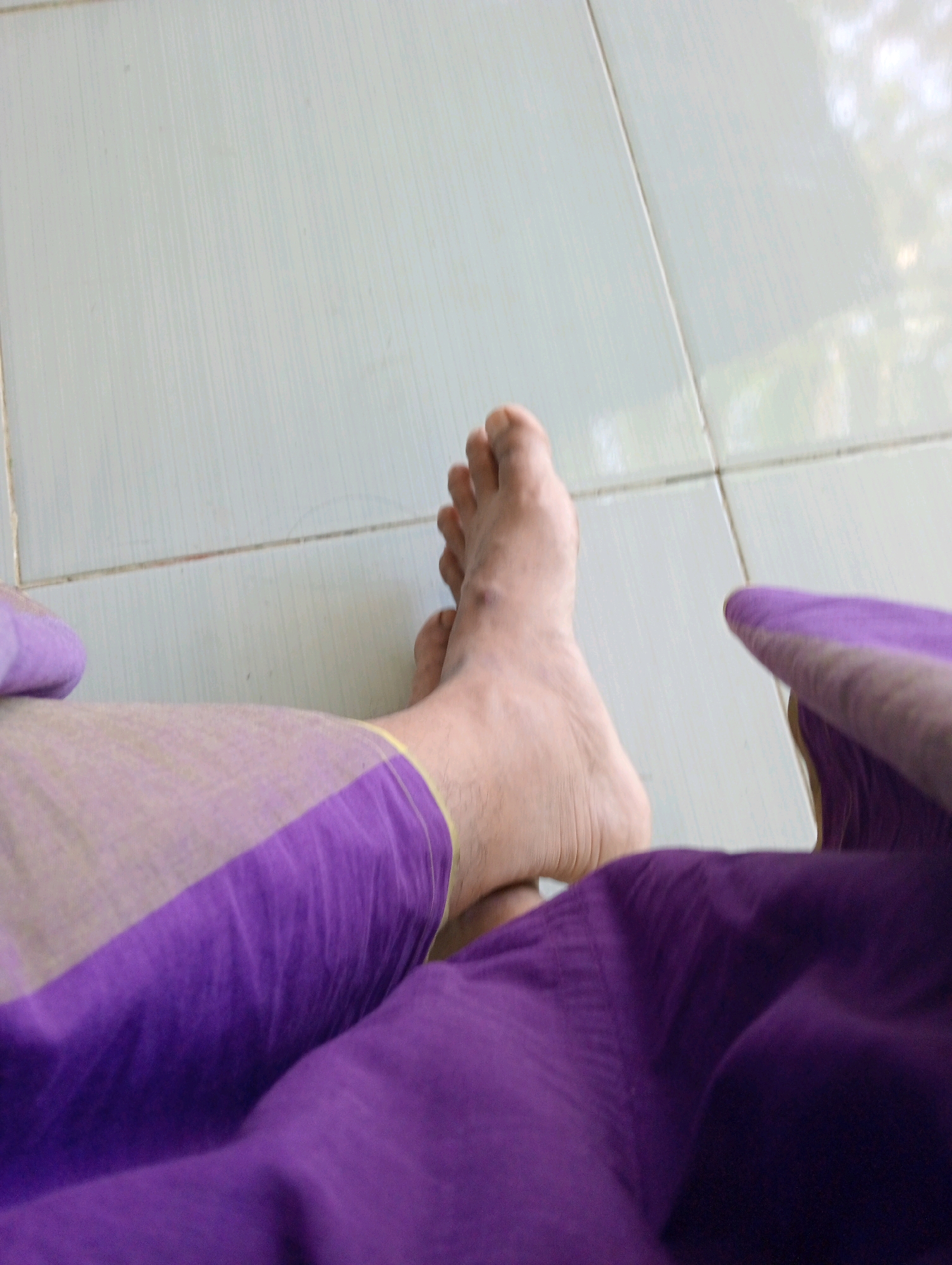 Two bare feet are visible on a tiled floor. One foot is positioned closer to the camera, with the toes slightly curled. The other foot is further away and is partially obscured by a piece of purple fabric. The fabric appears to be part of clothing, possibly a sarong or skirt, and has a lighter colored border. The floor is made of light-colored rectangular tiles with visible grout lines. The background shows reflections of greenery, suggesting an outdoor or near-outdoor environment. The scene does not provide enough information to determine a specific location in Pagerungan Kecil, Indonesia.