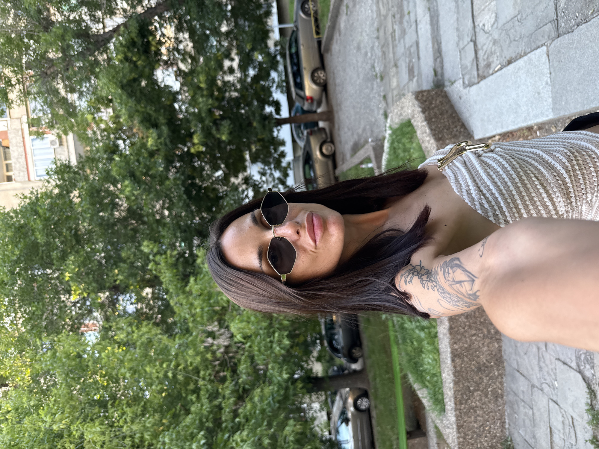 A woman with dark hair and sunglasses is taking a selfie outdoors. She is wearing a striped off-the-shoulder top with a gold clasp. A tattoo is visible on her left arm. Behind her, trees and a building can be seen, along with parked cars. The setting appears to be a residential courtyard in the city of Pleven, Bulgaria.